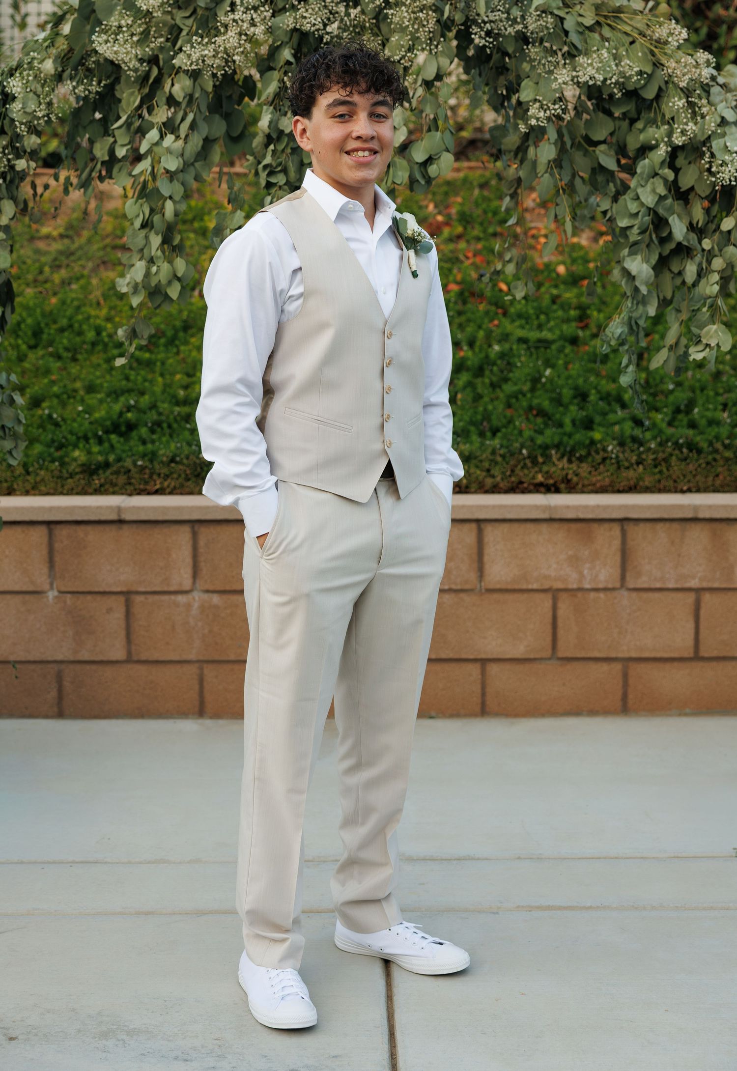 Young man in beige vest, white dress shirt, and matching trousers with white sneakers posing outdoors at a formal event.
