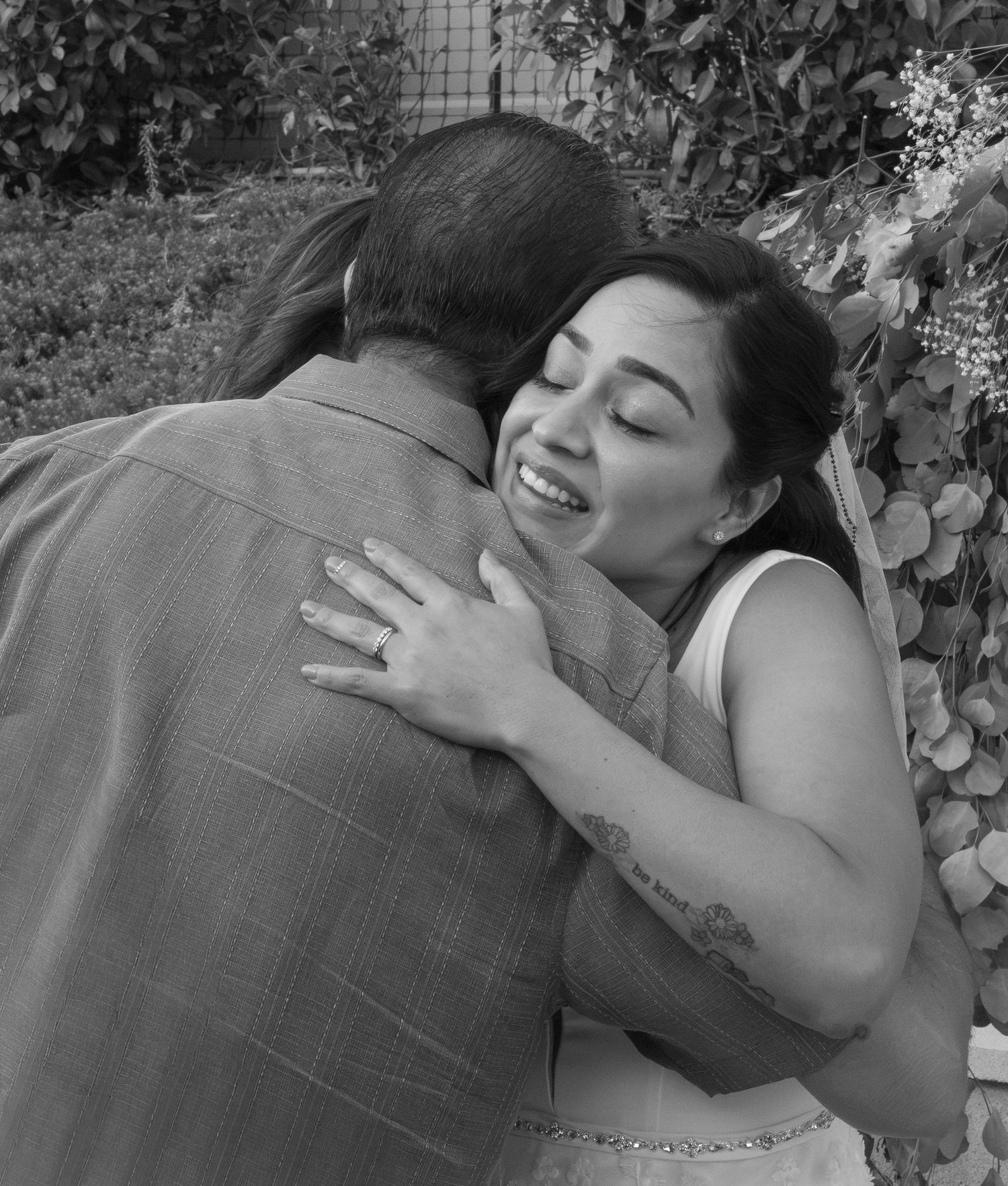 Black and white photo of a woman smiling with eyes closed, embracing a man outdoors near lush greenery.