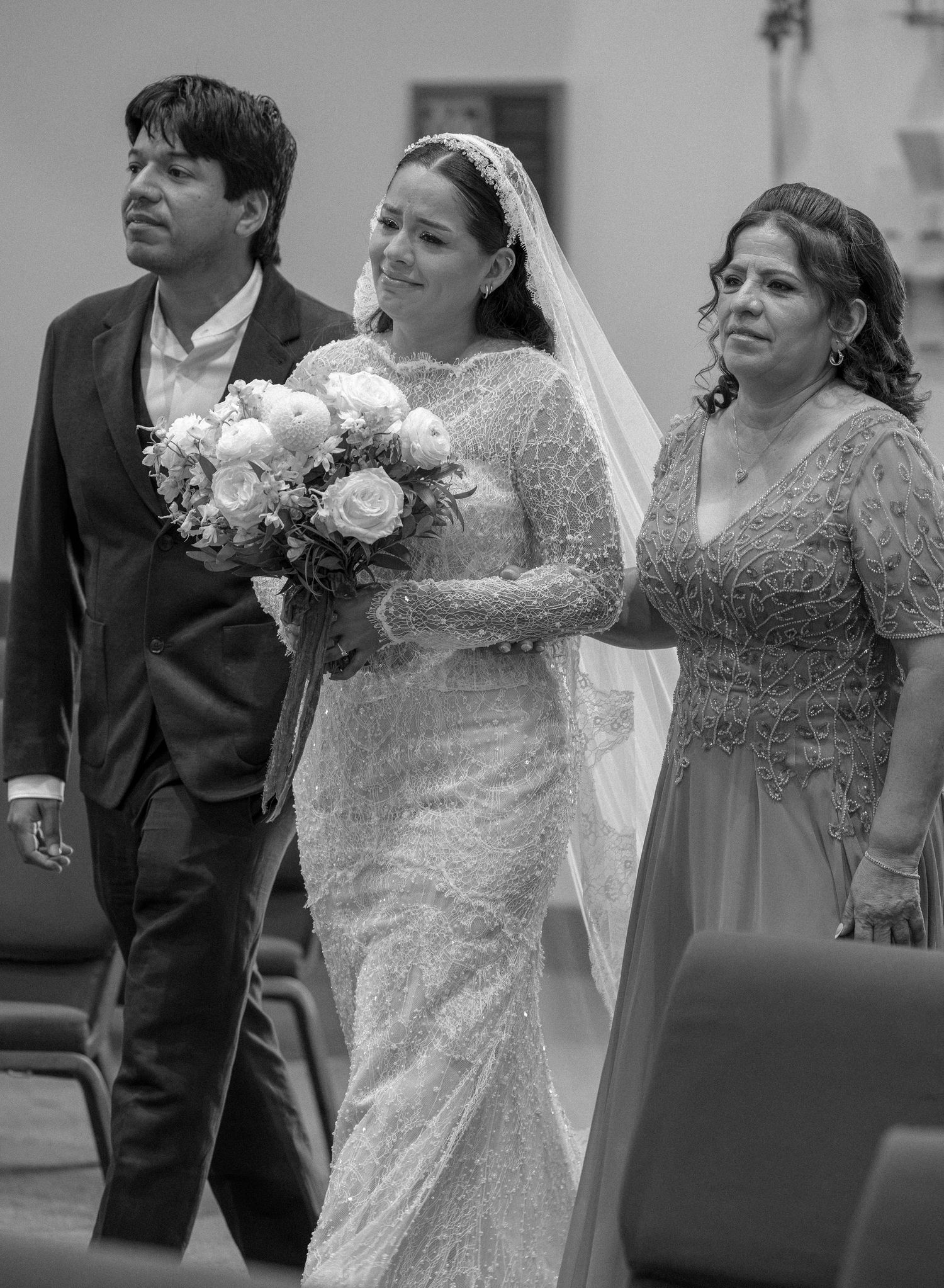 Black and white photo of a bride in a lace gown and veil holding a bouquet while walking down the aisle between two escorts.