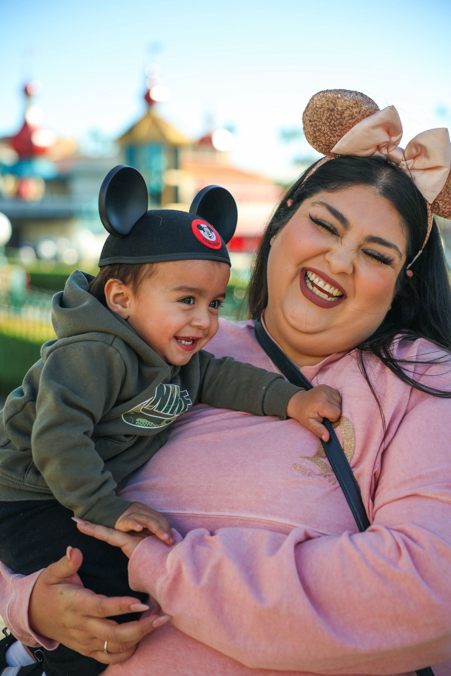 Laughing mother wearing Minnie Mouse ears holds smiling baby boy in Mickey Mouse hat at Disneyland.