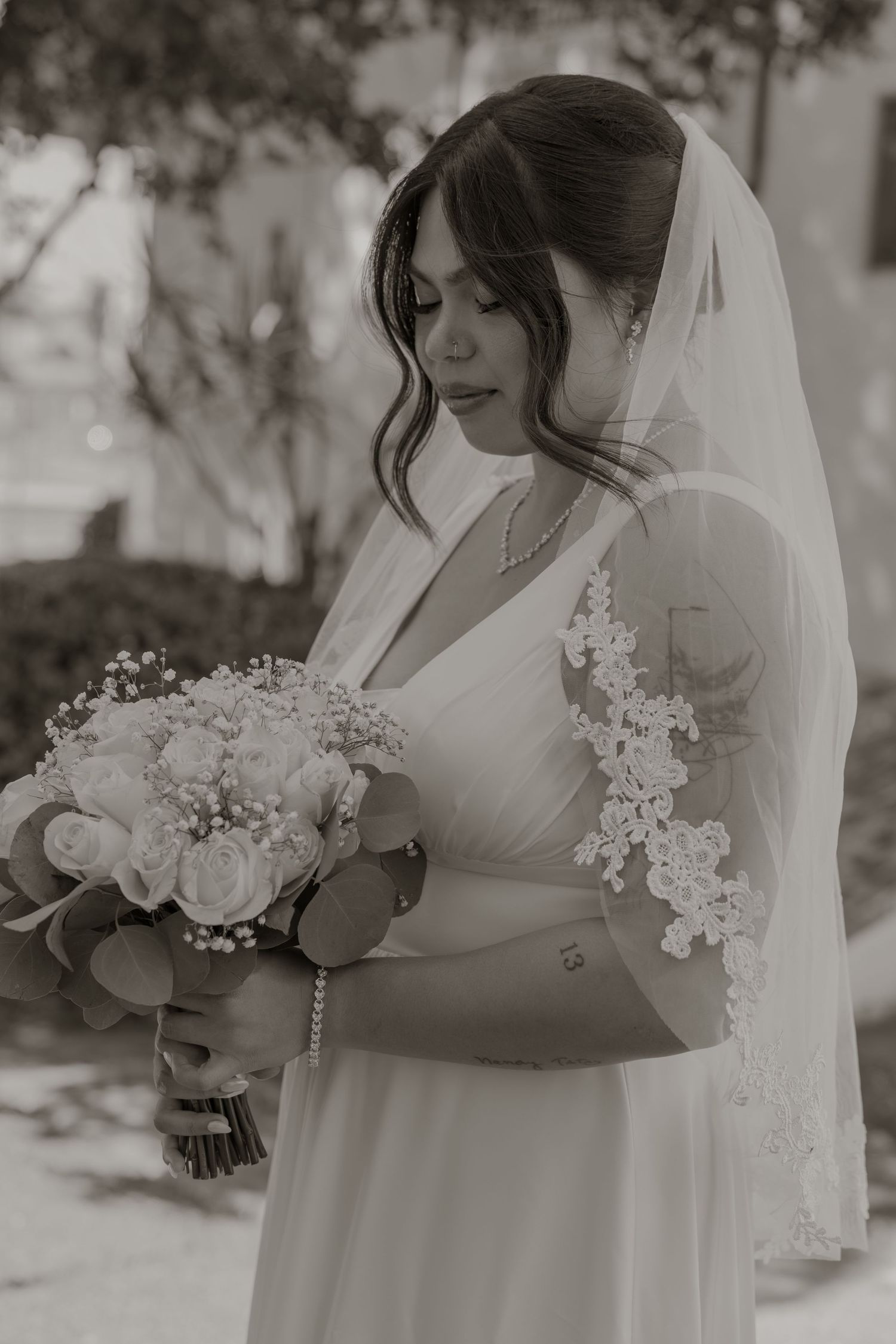 Elegant bride in white lace-trimmed veil gazes down at her white rose bouquet in black and white photo.