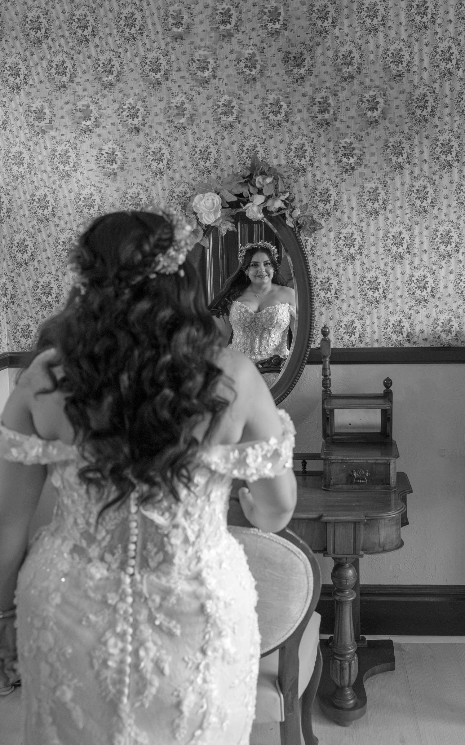 Bride in lace wedding dress views her reflection in vintage oval mirror, floral wallpaper background, black and white photo.