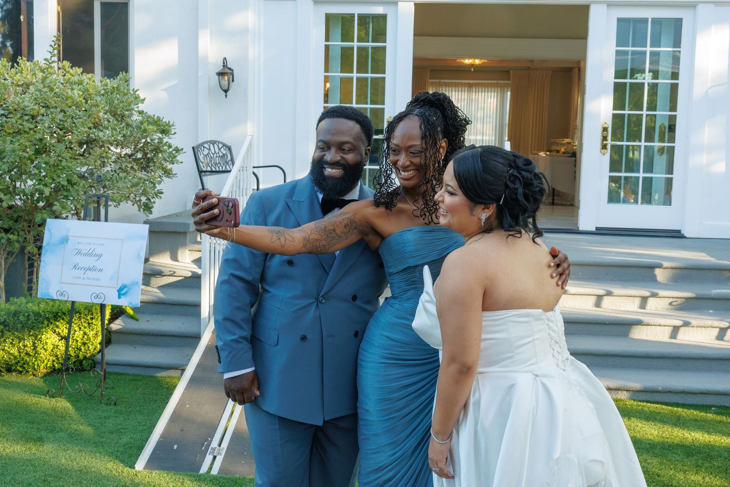 Wedding group selfie with groom in blue suit, bridesmaid in teal dress, and bride in white gown outdoors.