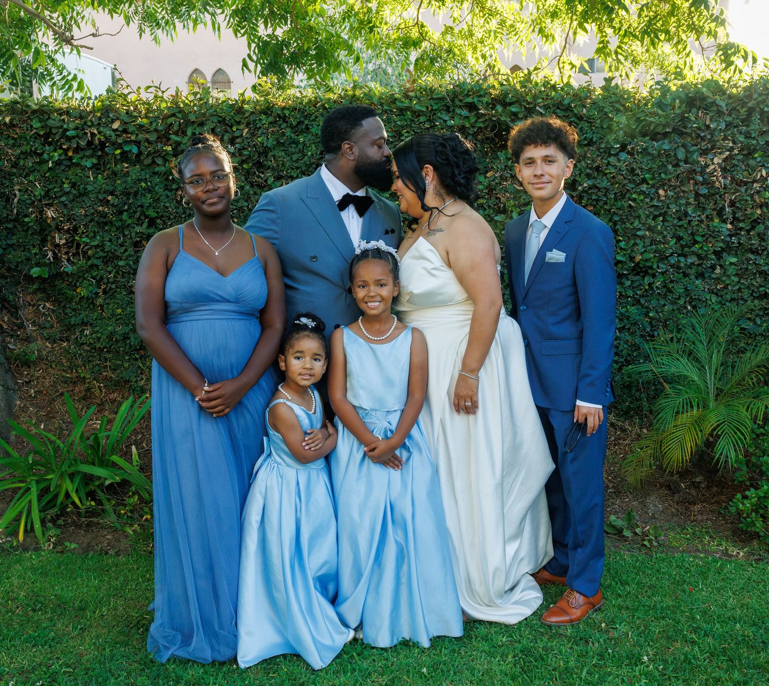 Wedding party group photo with bride, groom, and children in coordinating blue outfits in a garden.