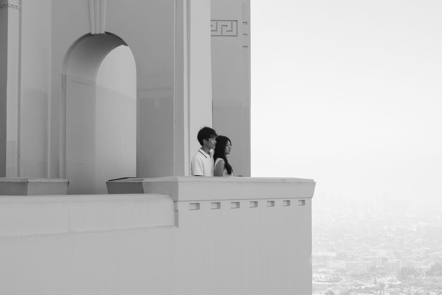 Romantic couple sharing a kiss on a white architectural balcony overlooking a misty ocean in black and white.