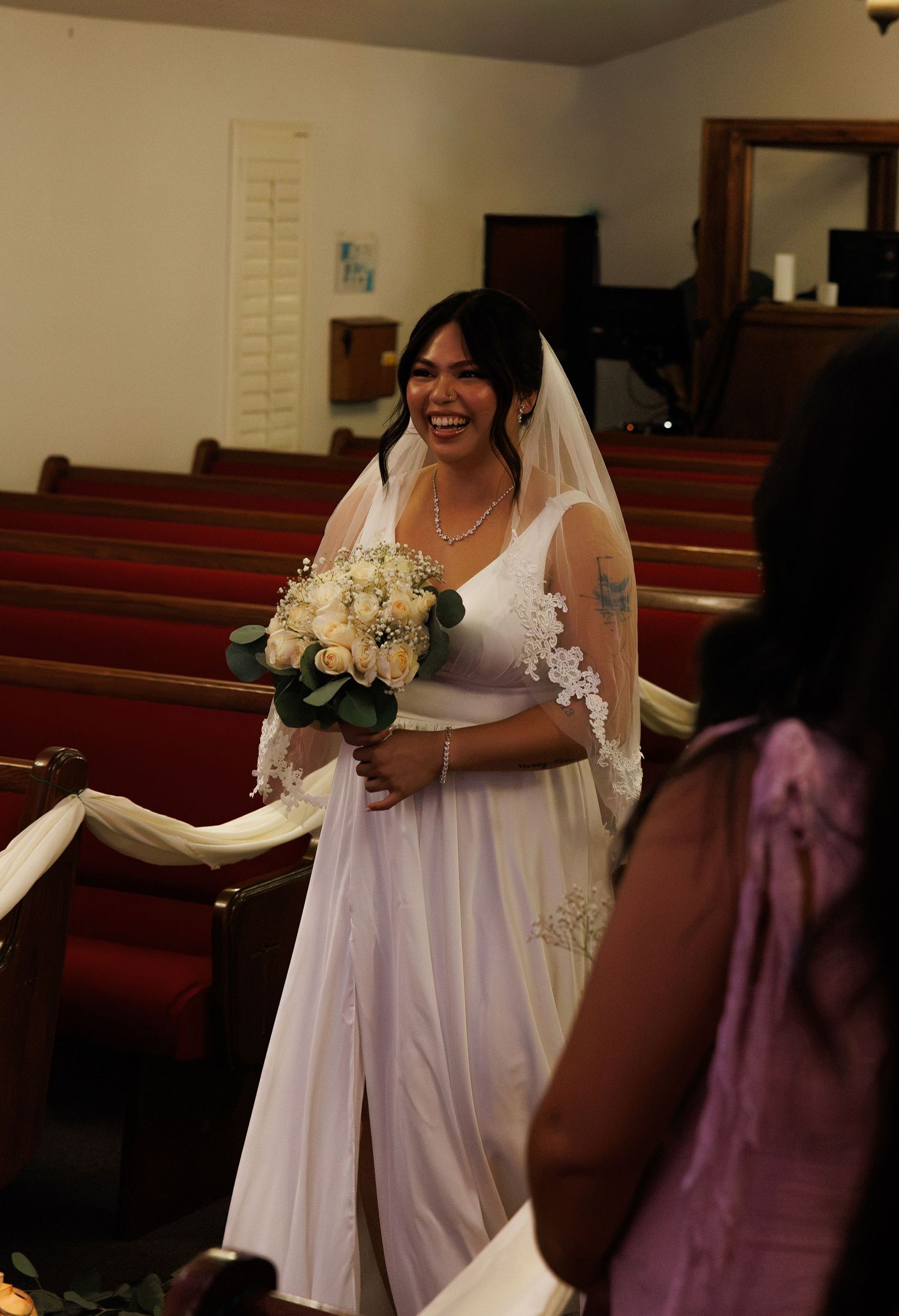 Smiling bride in white gown and lace veil walks down church aisle holding white rose bouquet.