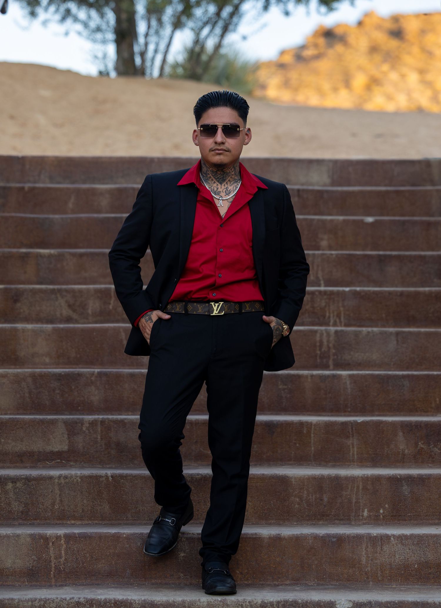 Man in black suit, red shirt, Louis Vuitton belt, and sunglasses poses on stone steps outdoors.
