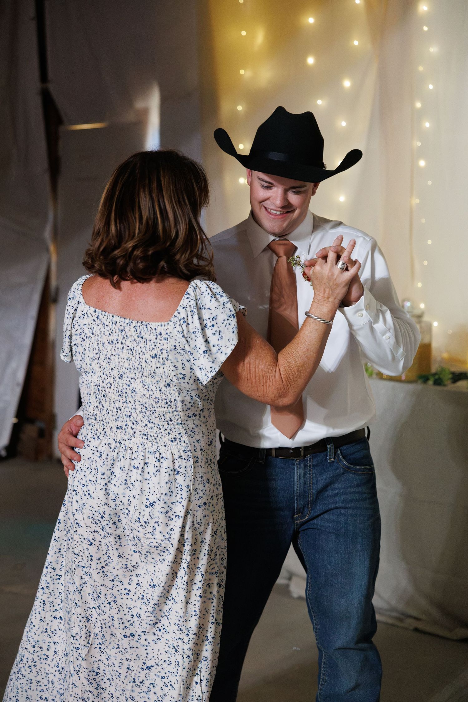 Smiling groom in black cowboy hat and peach tie dances with a woman in a floral dress at wedding reception.