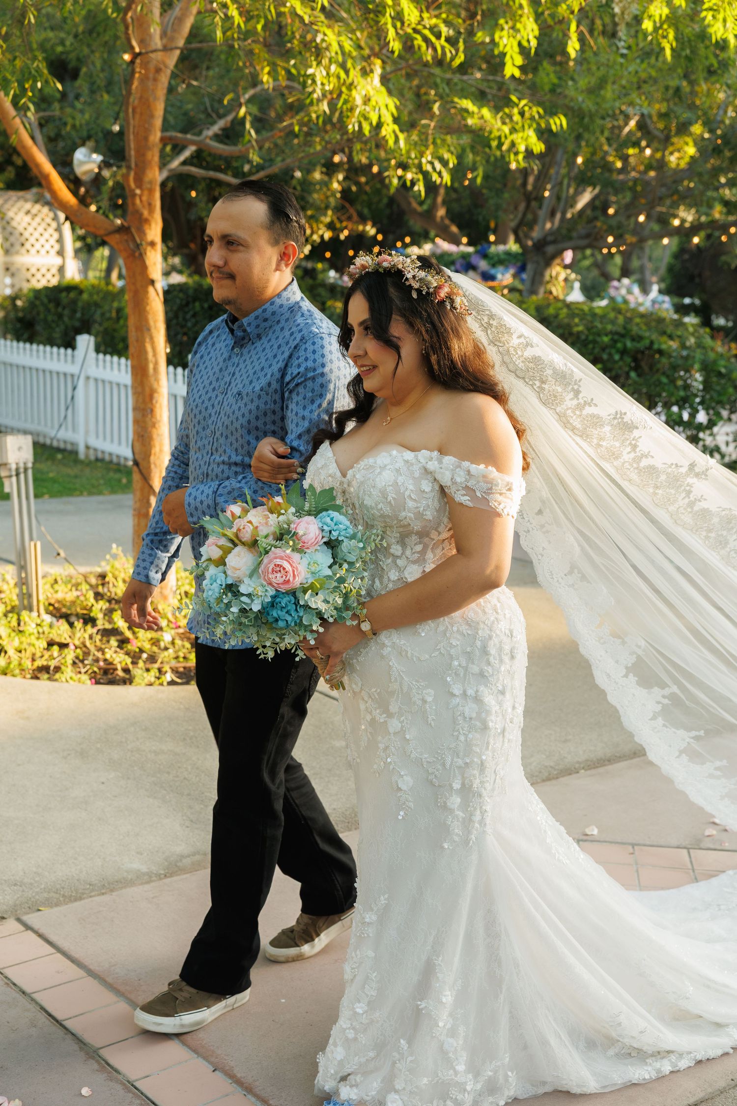 Bride in white lace gown holding floral bouquet walks with man in blue shirt through garden path.