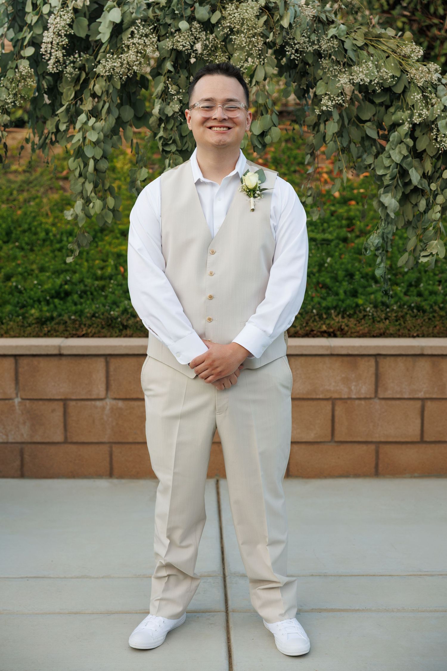Smiling groom in beige vest, white shirt, and boutonniere stands outdoors before lush greenery backdrop.