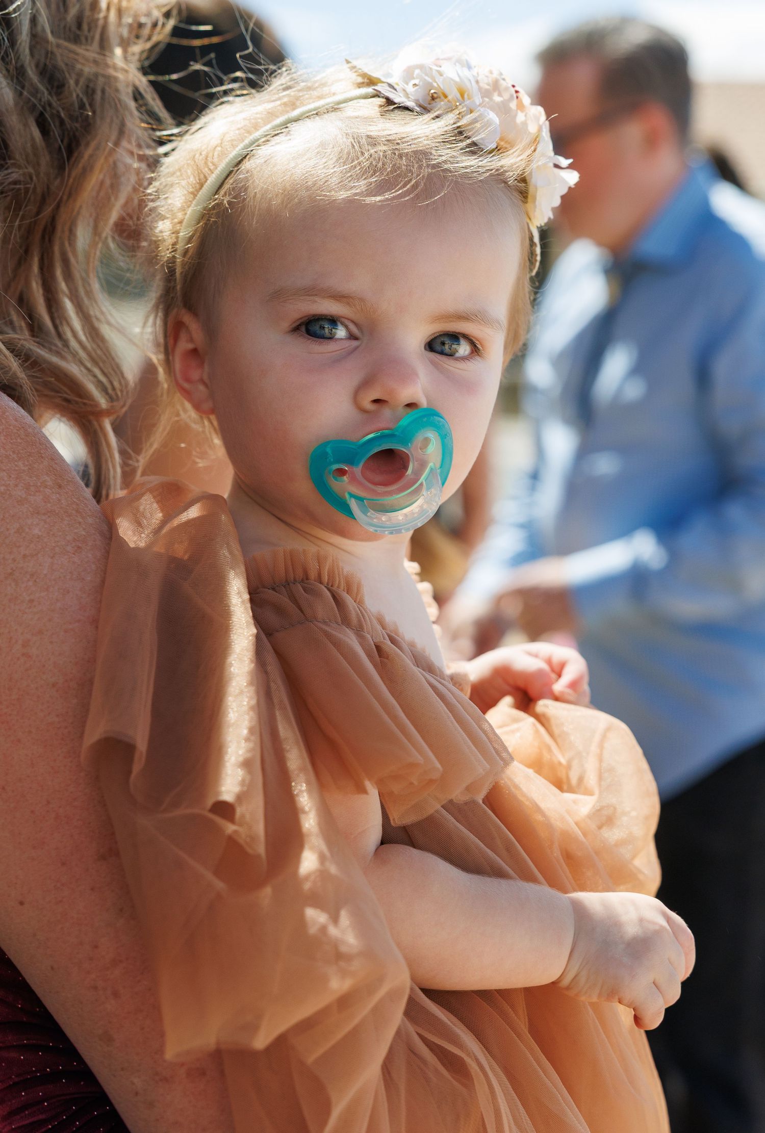 Adorable toddler girl in peach tulle dress with floral hair accessory and teal pacifier being held at outdoor wedding.