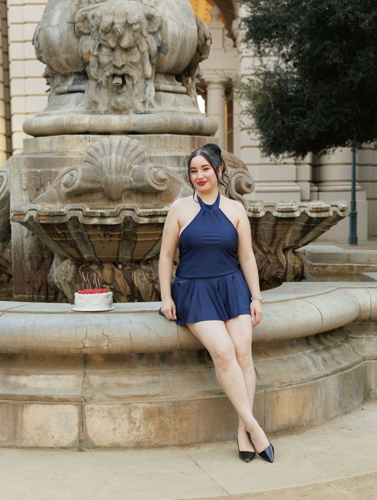 Woman in navy blue halter romper sitting on ornate stone fountain ledge with a cake beside her.