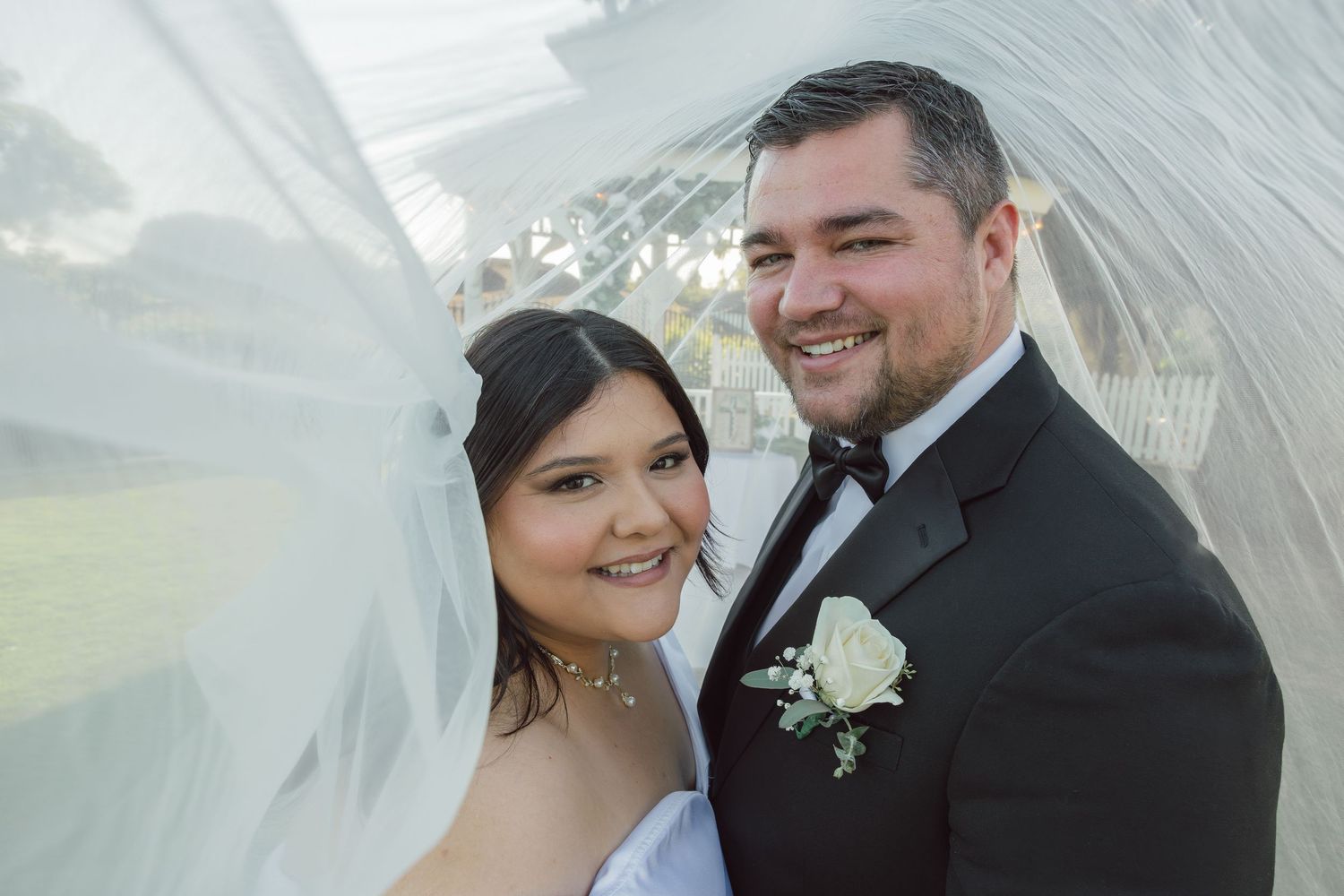 A smiling bride and groom pose under a flowing white veil on their wedding day, with the groom in a tuxedo.