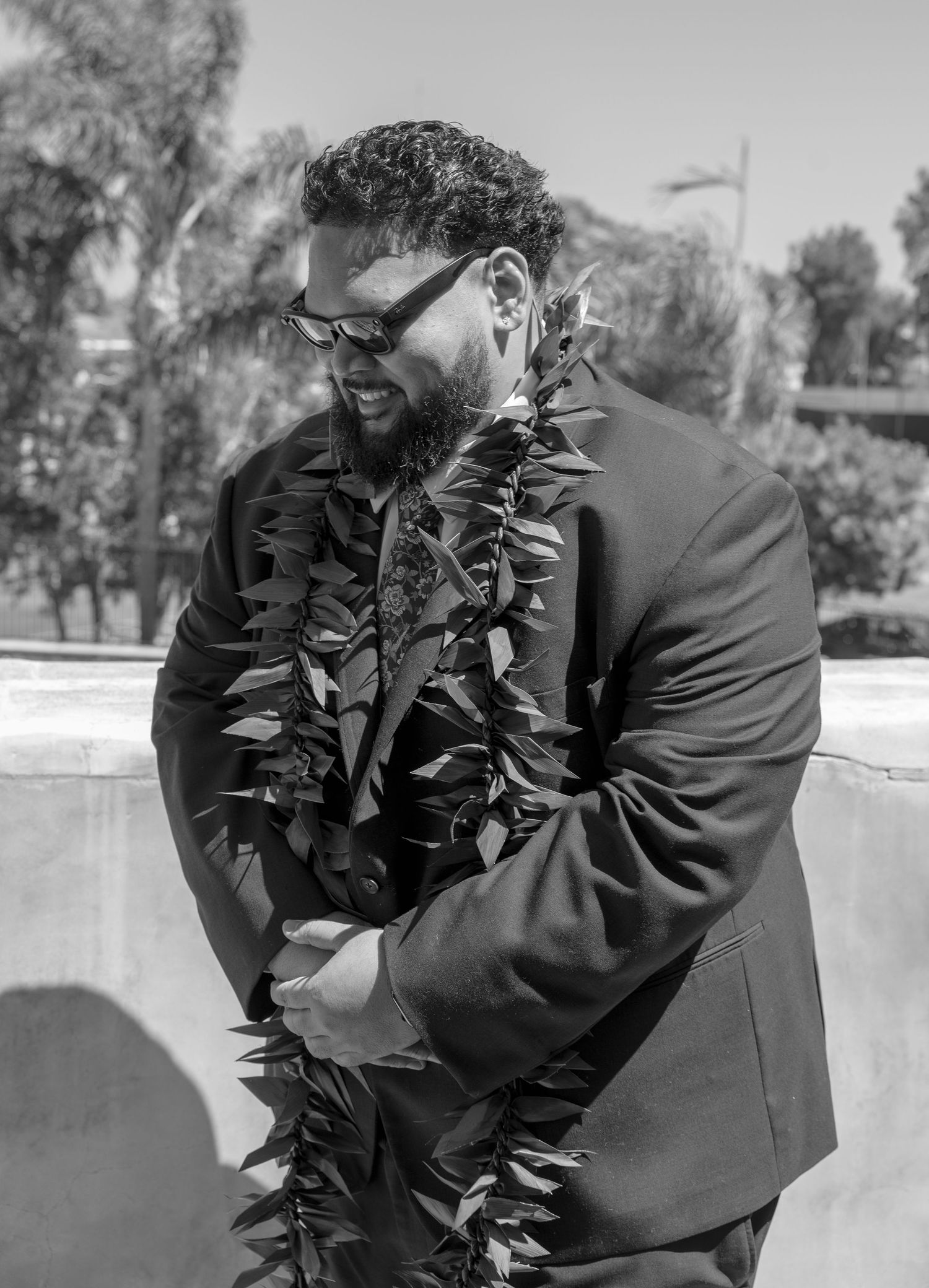 Bearded groom in black suit wearing floral lei and sunglasses smiling at outdoor wedding.