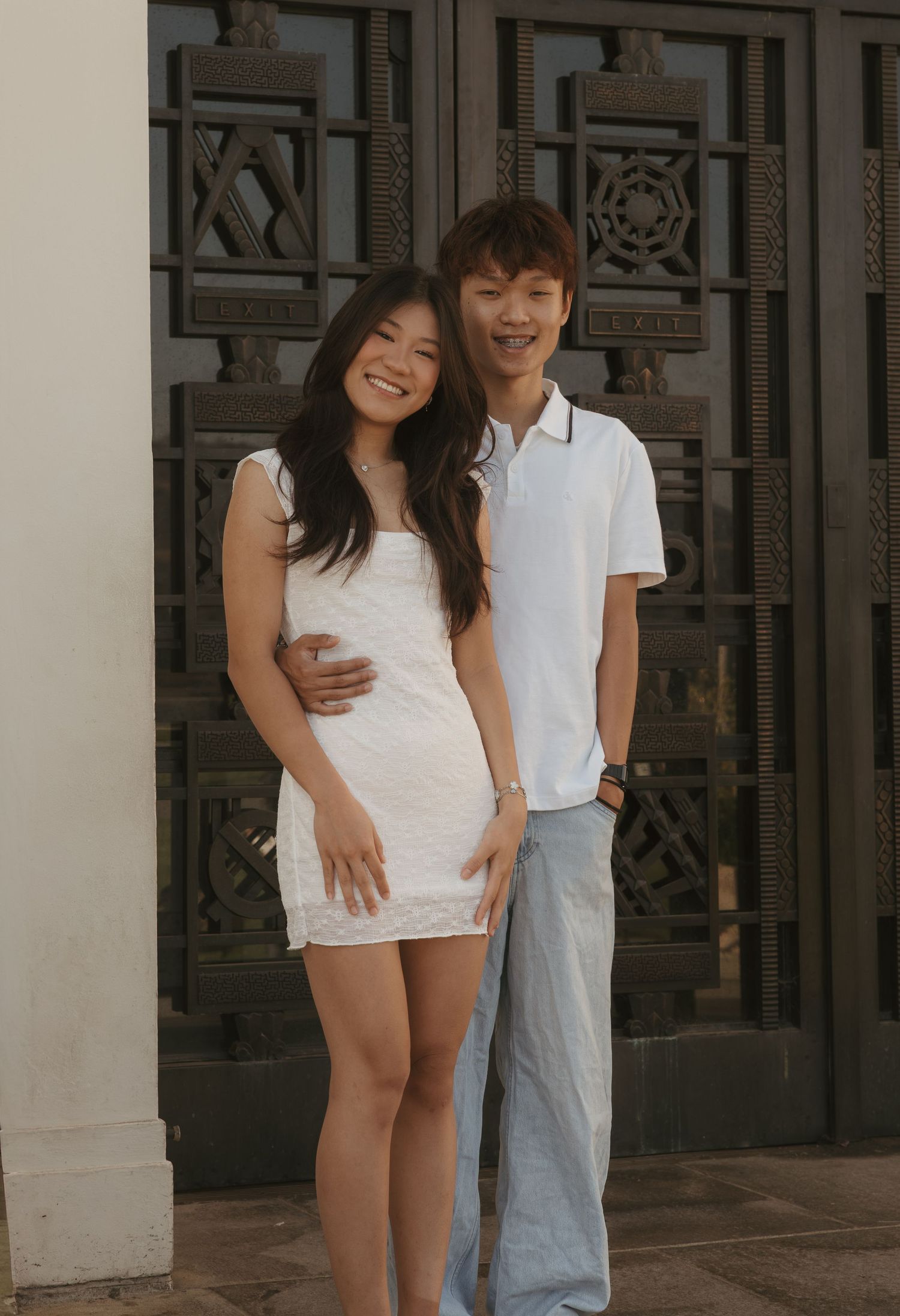 A young couple poses smiling in front of ornate dark doors, wearing white outfits on a sunny day.
