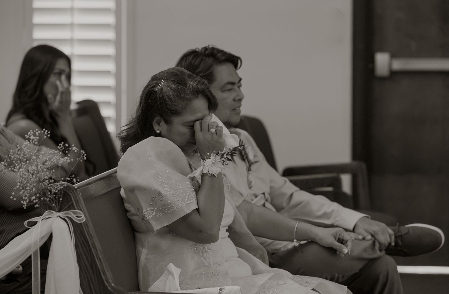 Black and white photo of emotional couple in wedding attire seated together, bride wiping tears, guests in background.