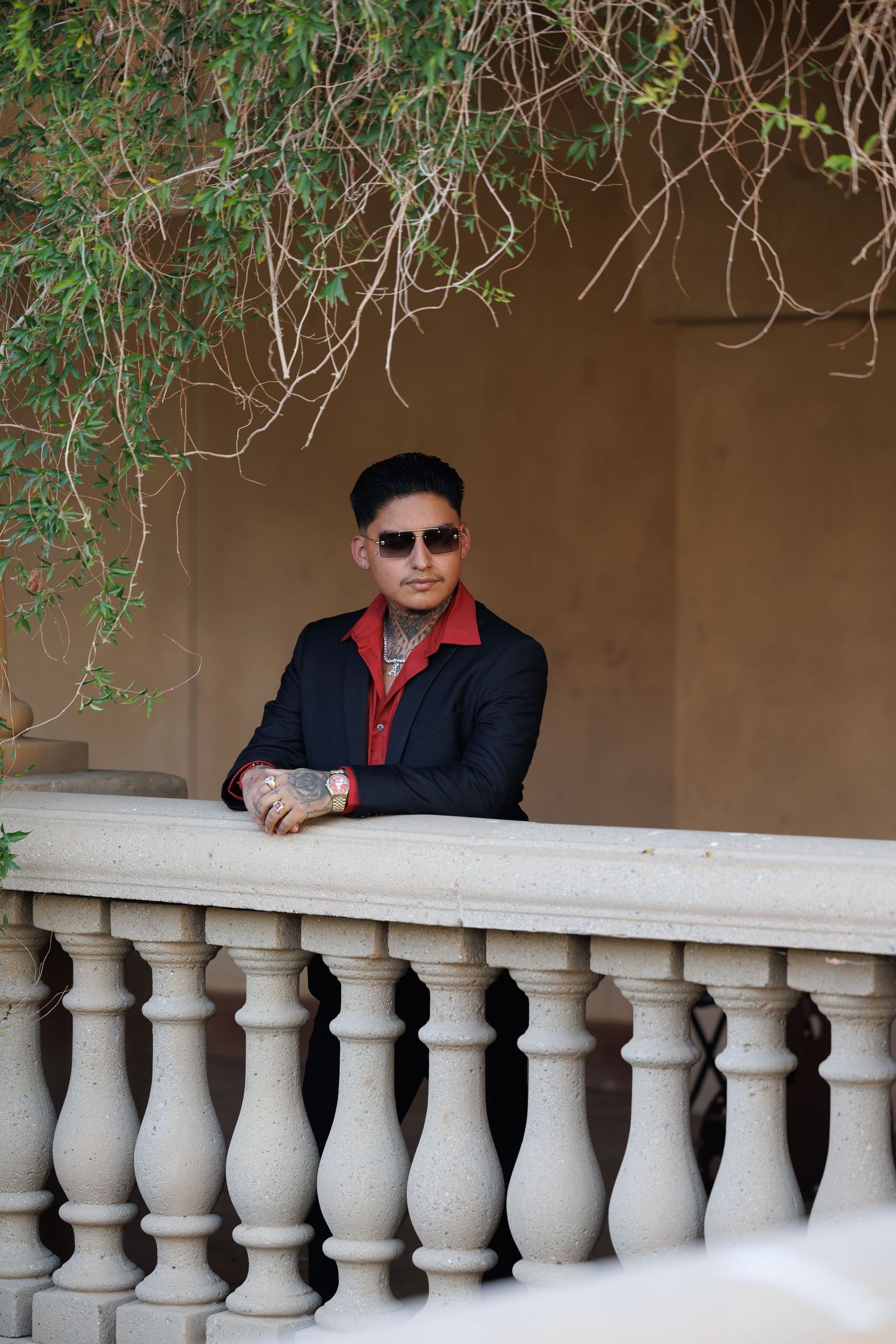 A tattooed man in a black and red jacket and sunglasses leans on a stone balustrade balcony surrounded by greenery.