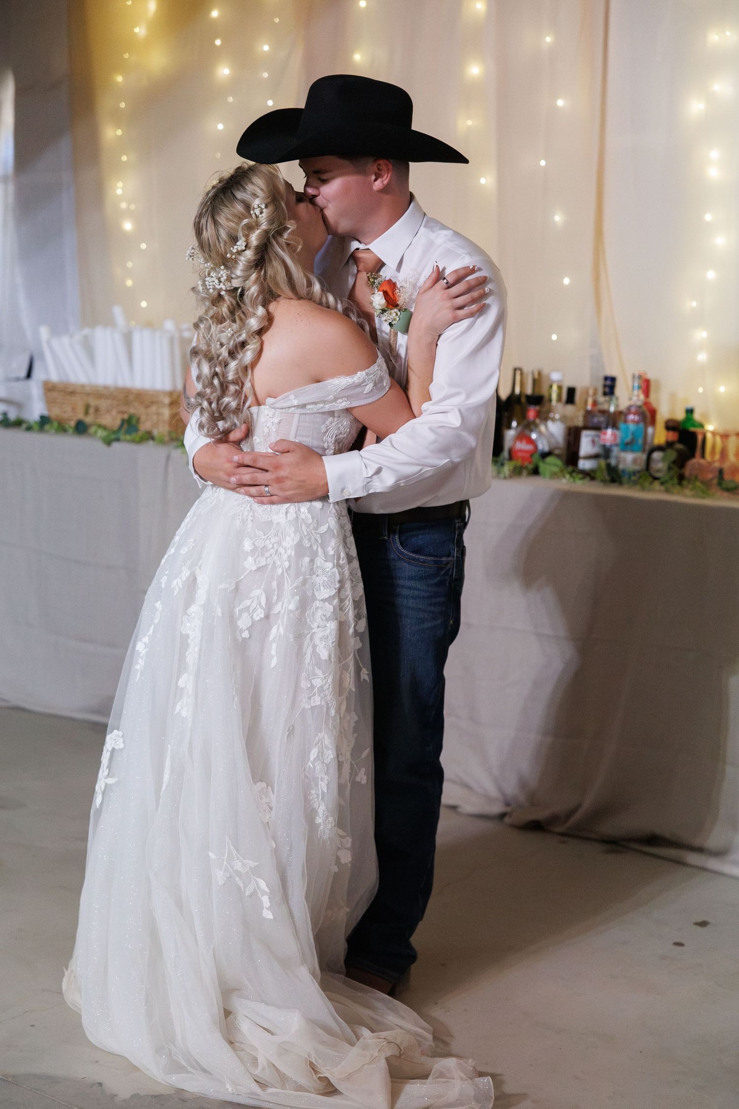 Bride and groom share a romantic kiss during their first dance, groom wearing a black cowboy hat at wedding reception.