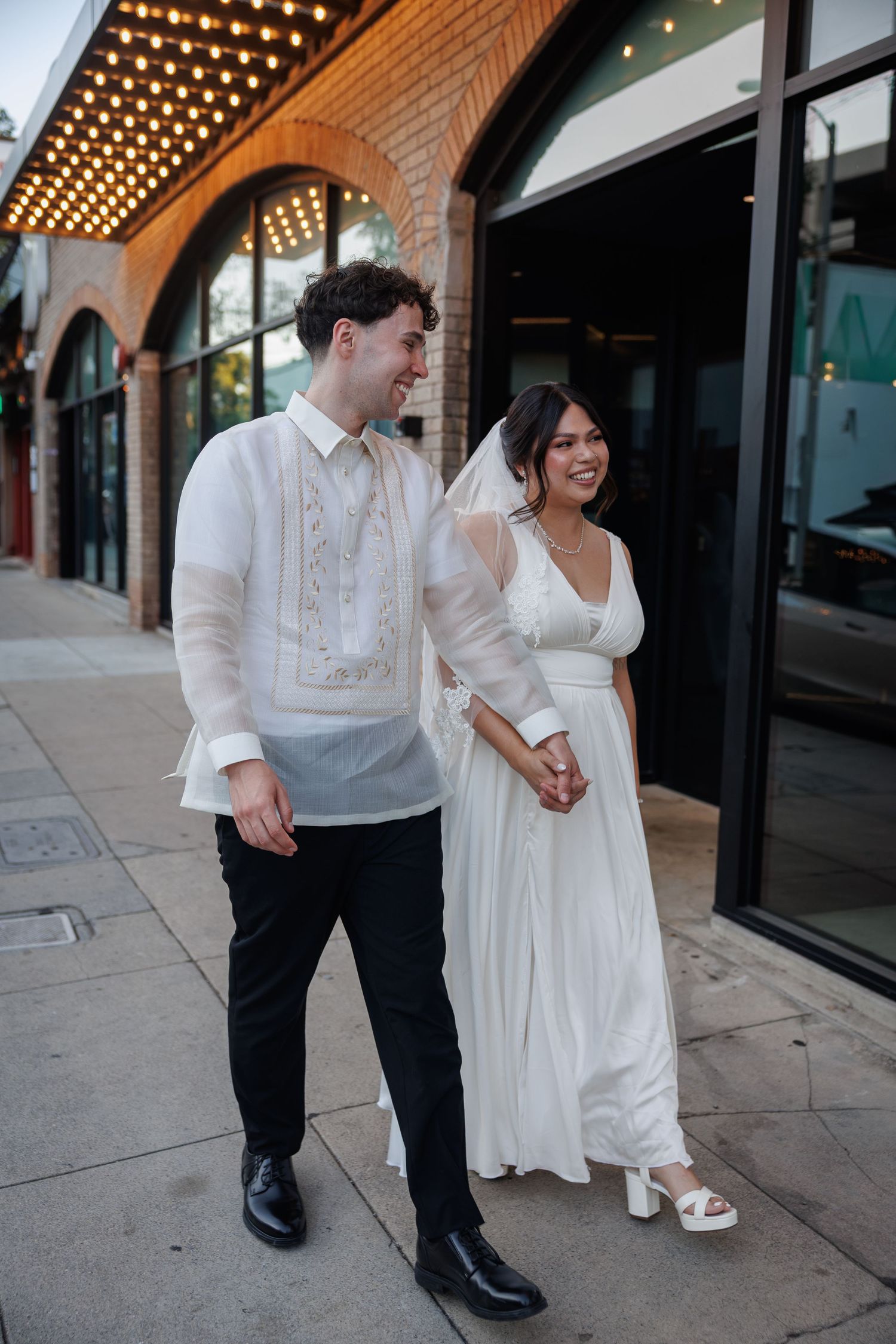 Couple in Filipino barong and white bridal gown holding hands outside a venue with marquee lights.