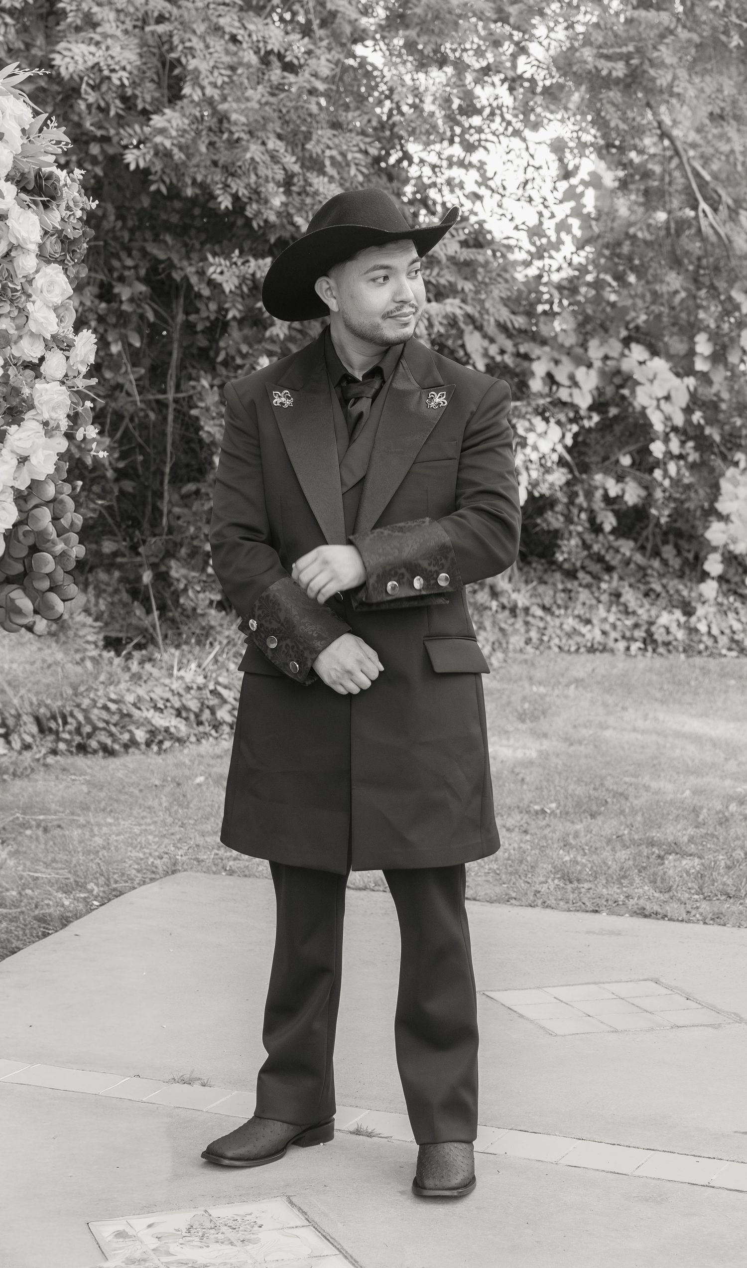 A man in a black cowboy hat and long coat stands outdoors in a black and white photo near blooming flowers.