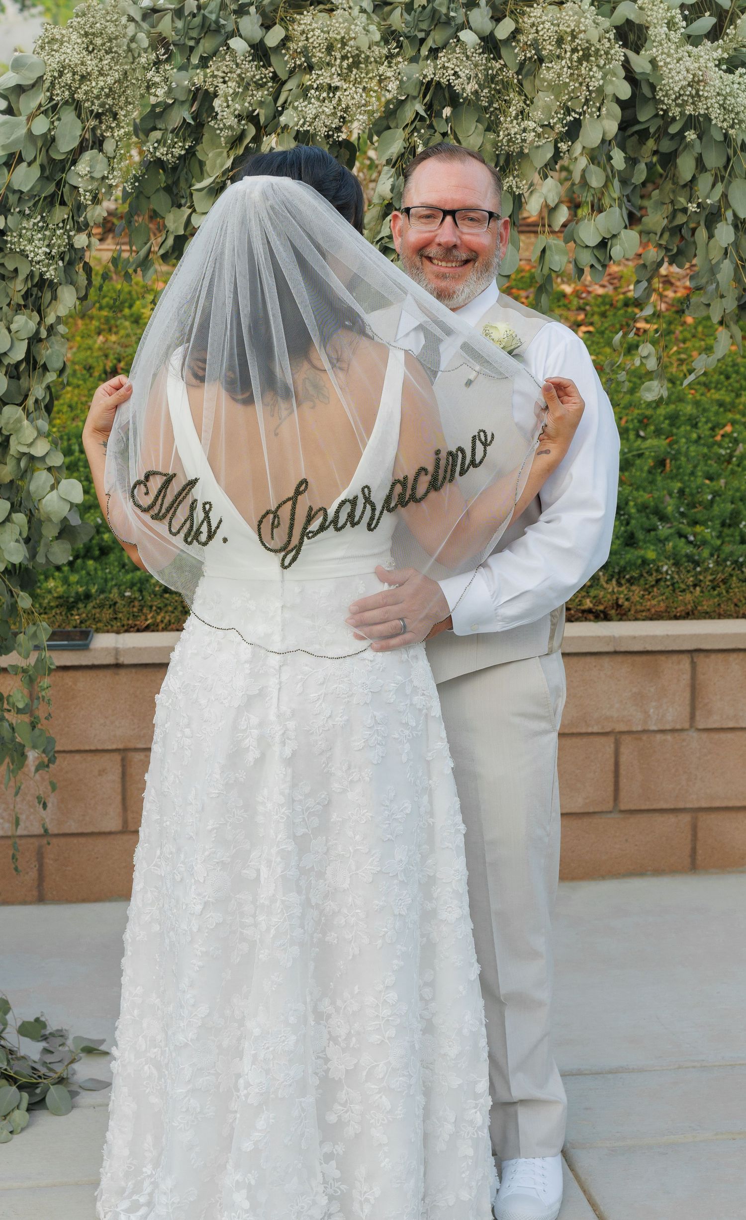 Bride in lace wedding dress with Mrs. Sparacino veil held by smiling groom in tan suit outdoors.