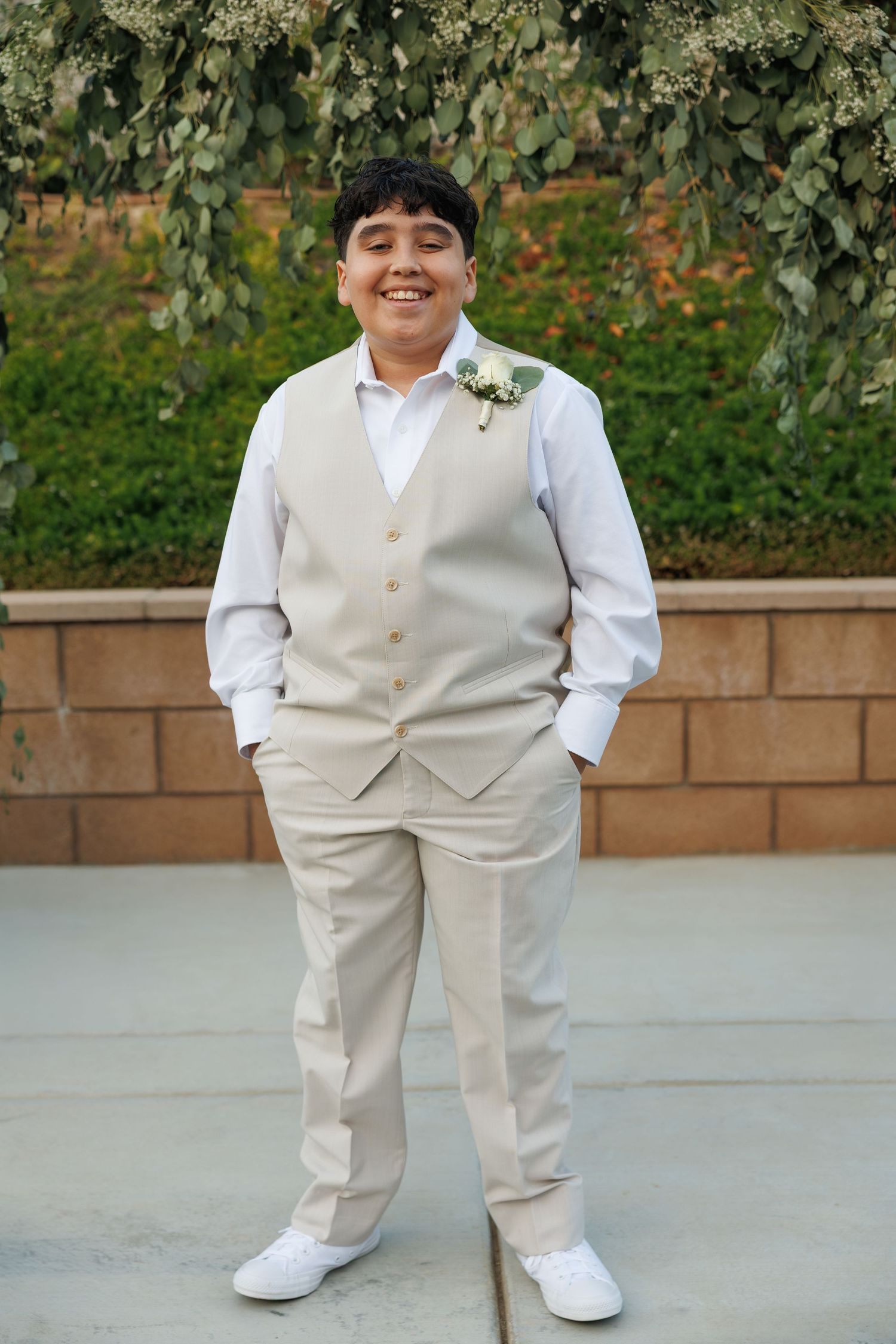 Smiling boy in beige vest suit with boutonniere stands outdoors at a wedding ceremony.