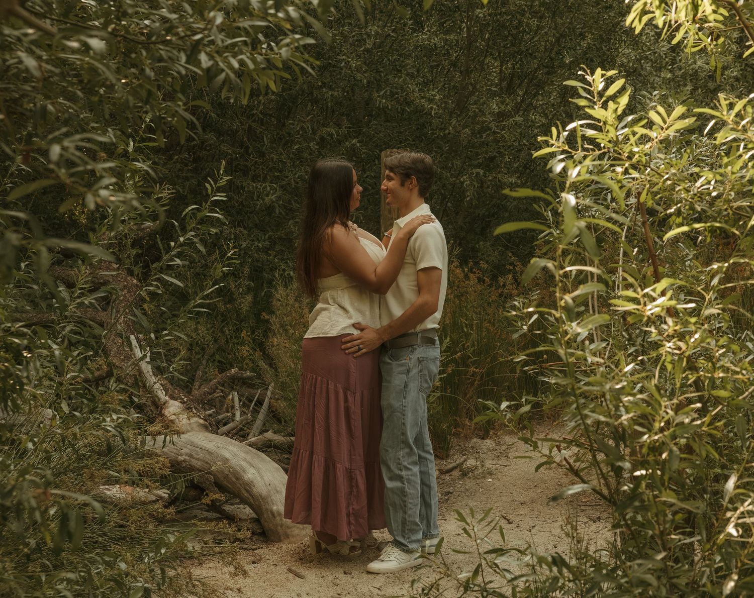 A couple embraces romantically in a lush green forest clearing, surrounded by trees and foliage.