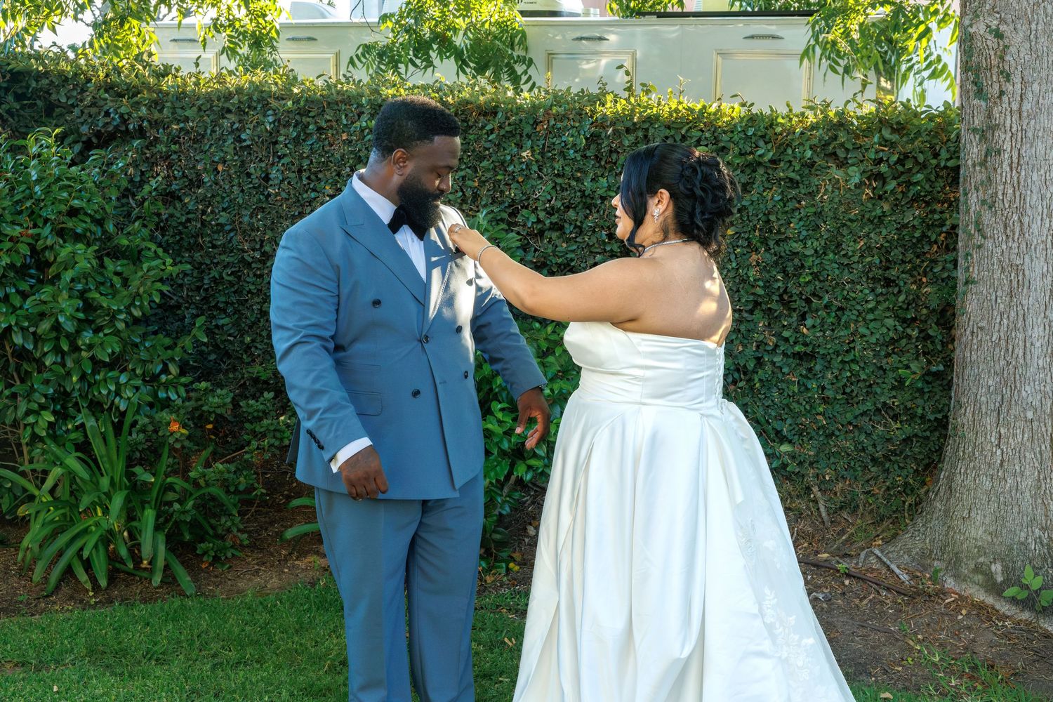 Bride in white gown adjusting groom's boutonniere before their wedding ceremony outdoors.
