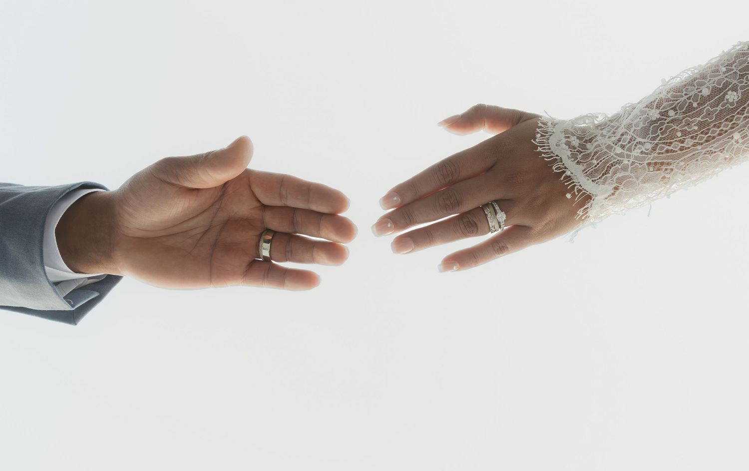 Bride and groom reaching hands toward each other, both wearing wedding rings, with a lace-cuffed sleeve against a bright white background.