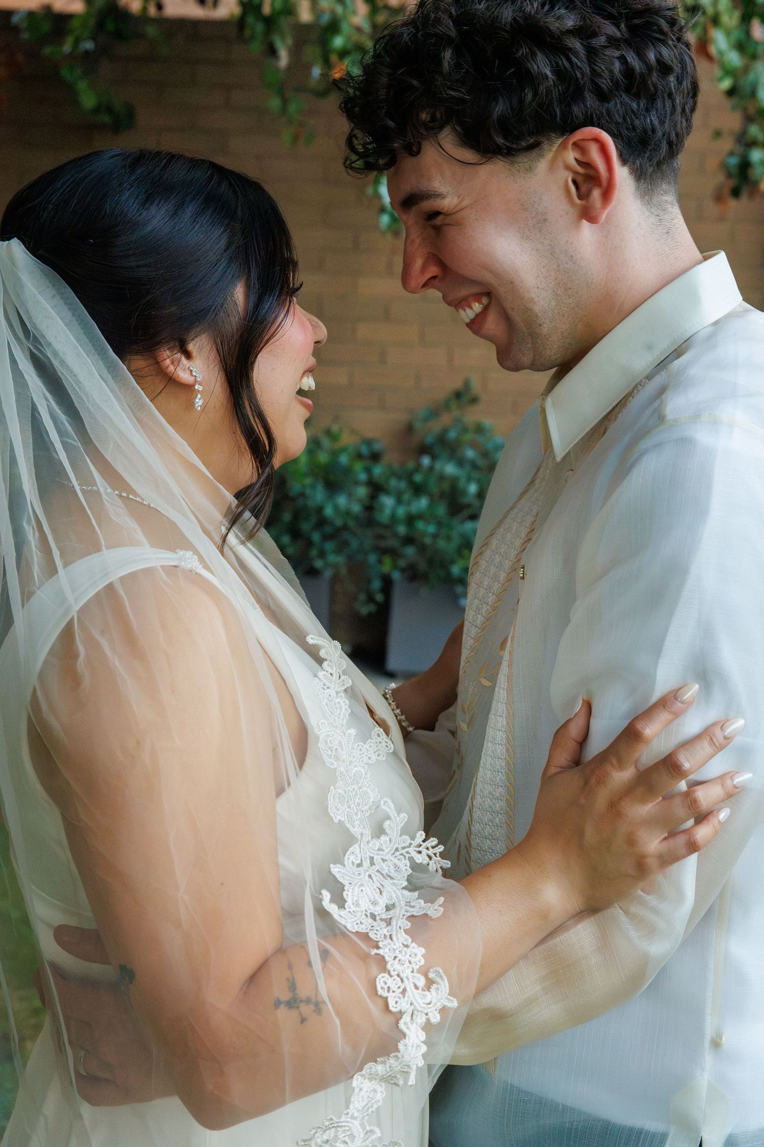 A bride and groom share a joyful moment, smiling at each other, bride in lace veil and groom in cream barong tagalog.