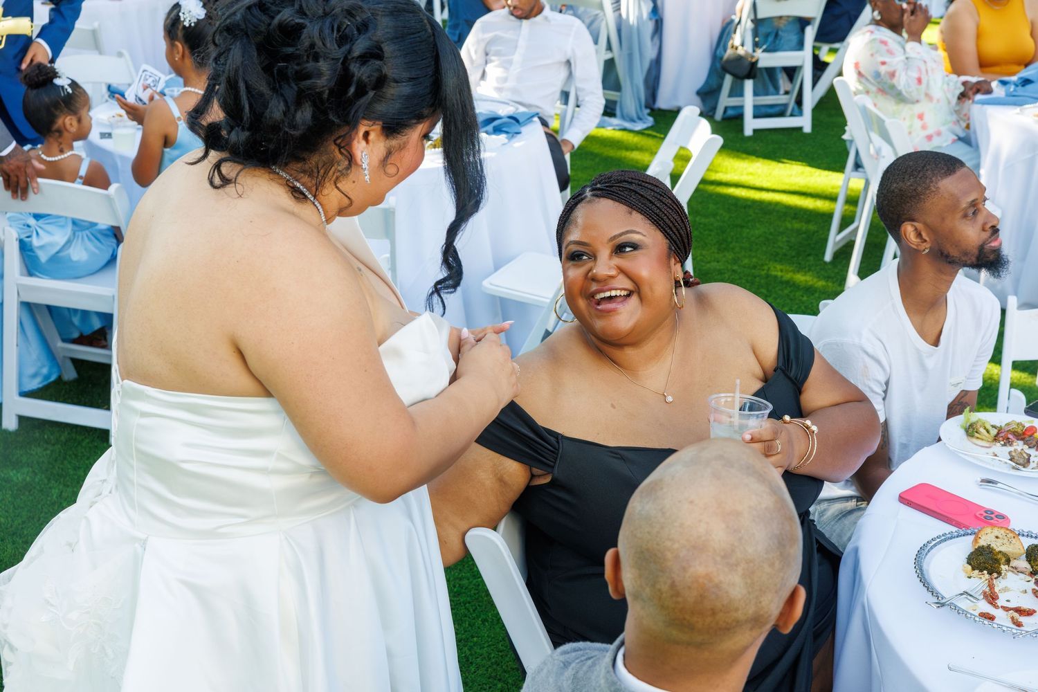 Bride in white gown greets smiling guest at outdoor wedding reception on green lawn.