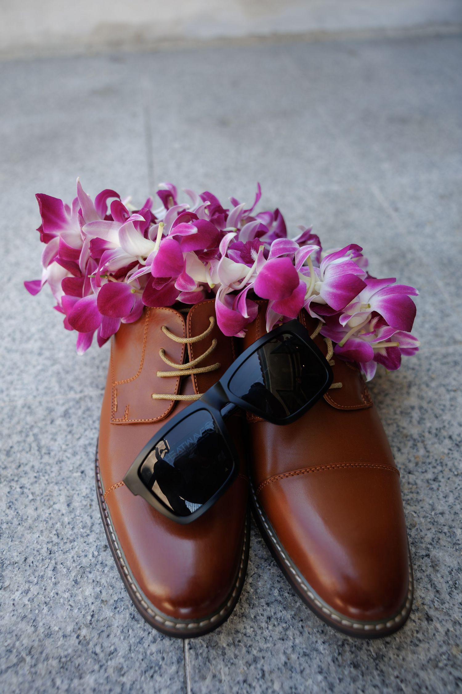 Brown leather dress shoes adorned with purple orchid lei and black sunglasses on gray stone surface.
