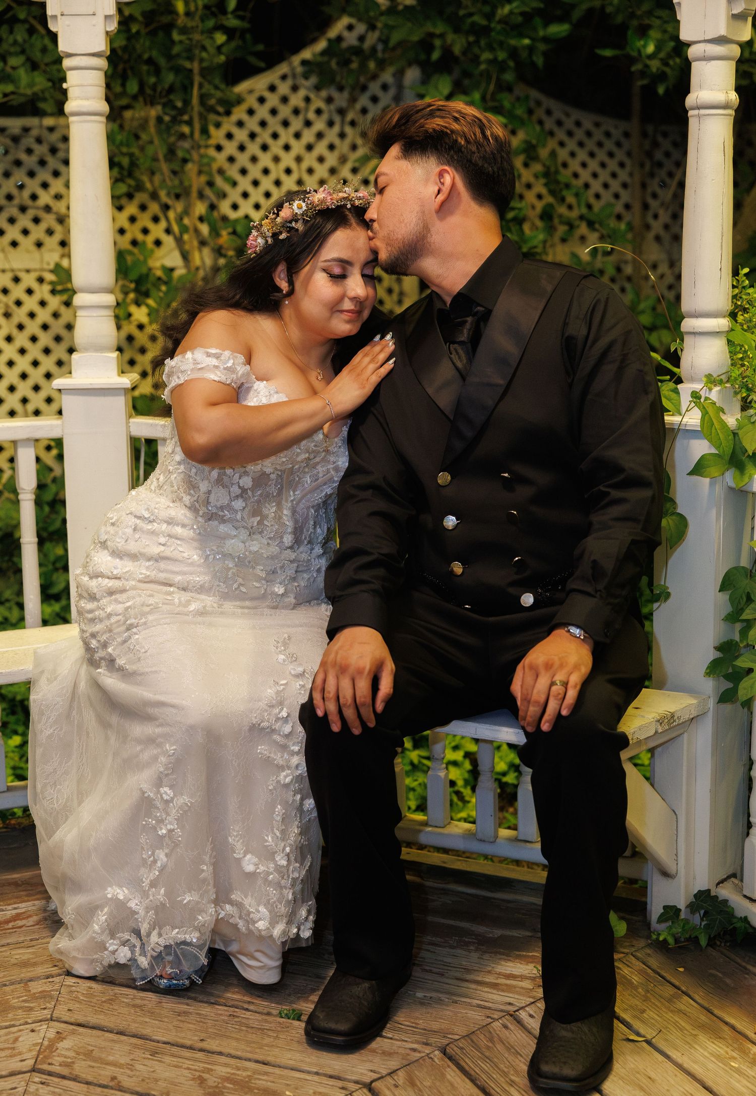 Bride and groom share a romantic kiss on a charming gazebo bench surrounded by lush greenery.