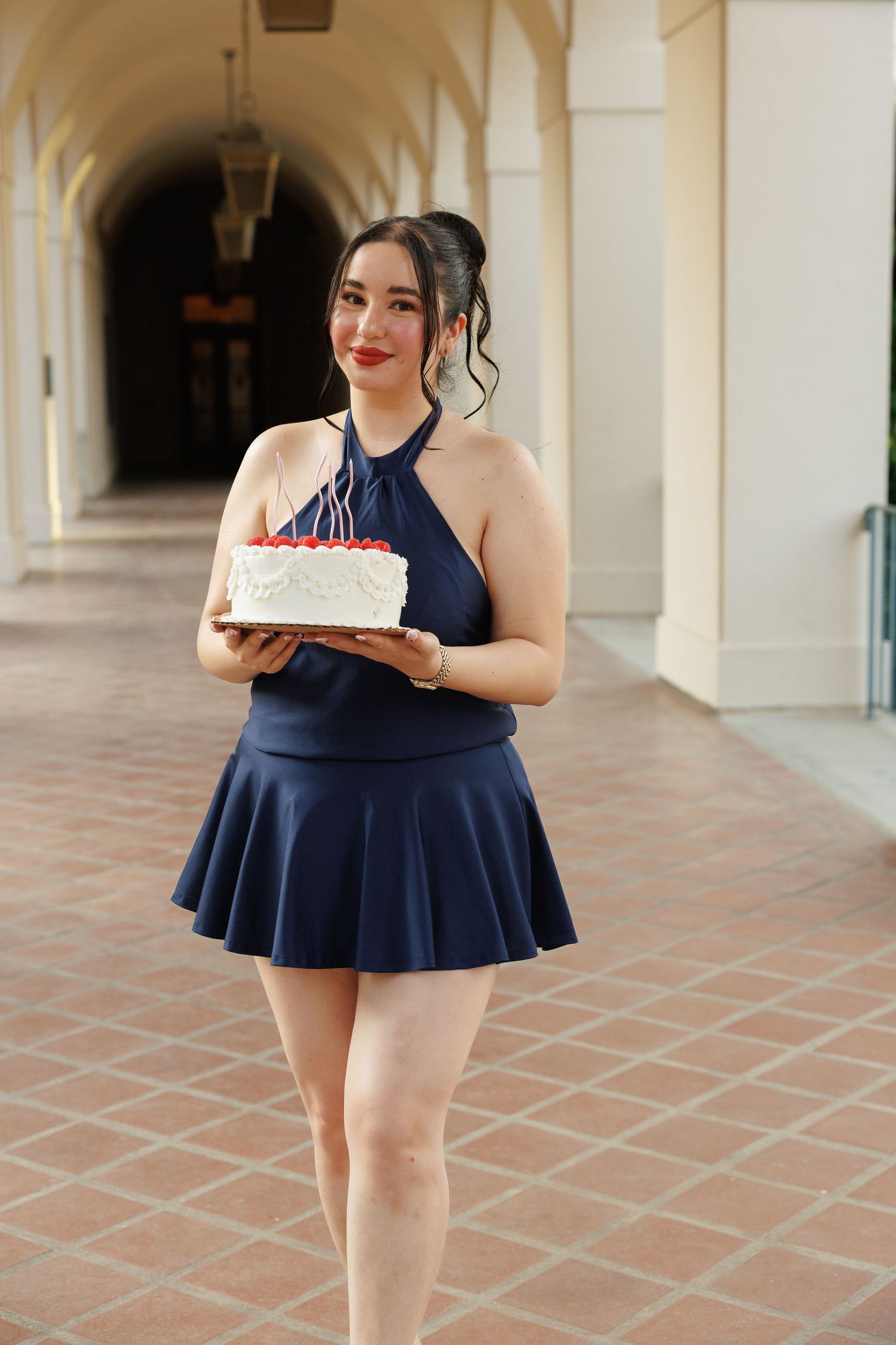 Young woman in navy blue halter dress holding a white birthday cake with candles in an elegant arched corridor.