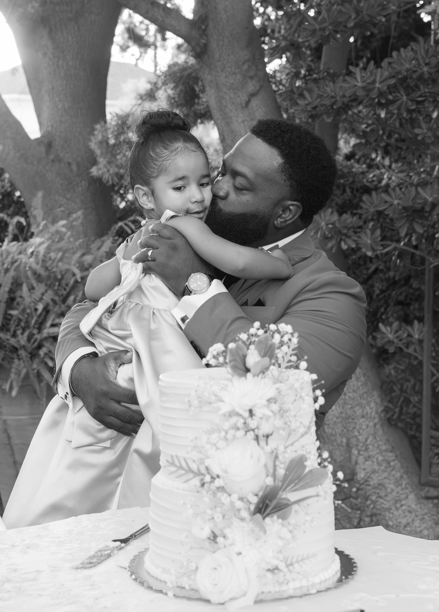 Groom in gray suit kissing young child near wedding cake in black and white photo.