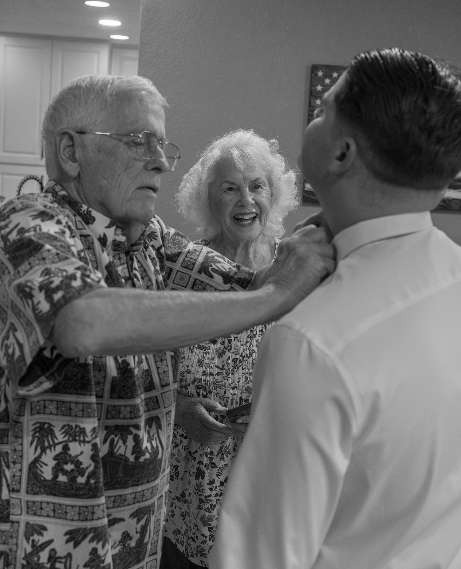 Elderly man in Hawaiian shirt adjusting groom's collar while smiling woman watches at wedding reception.
