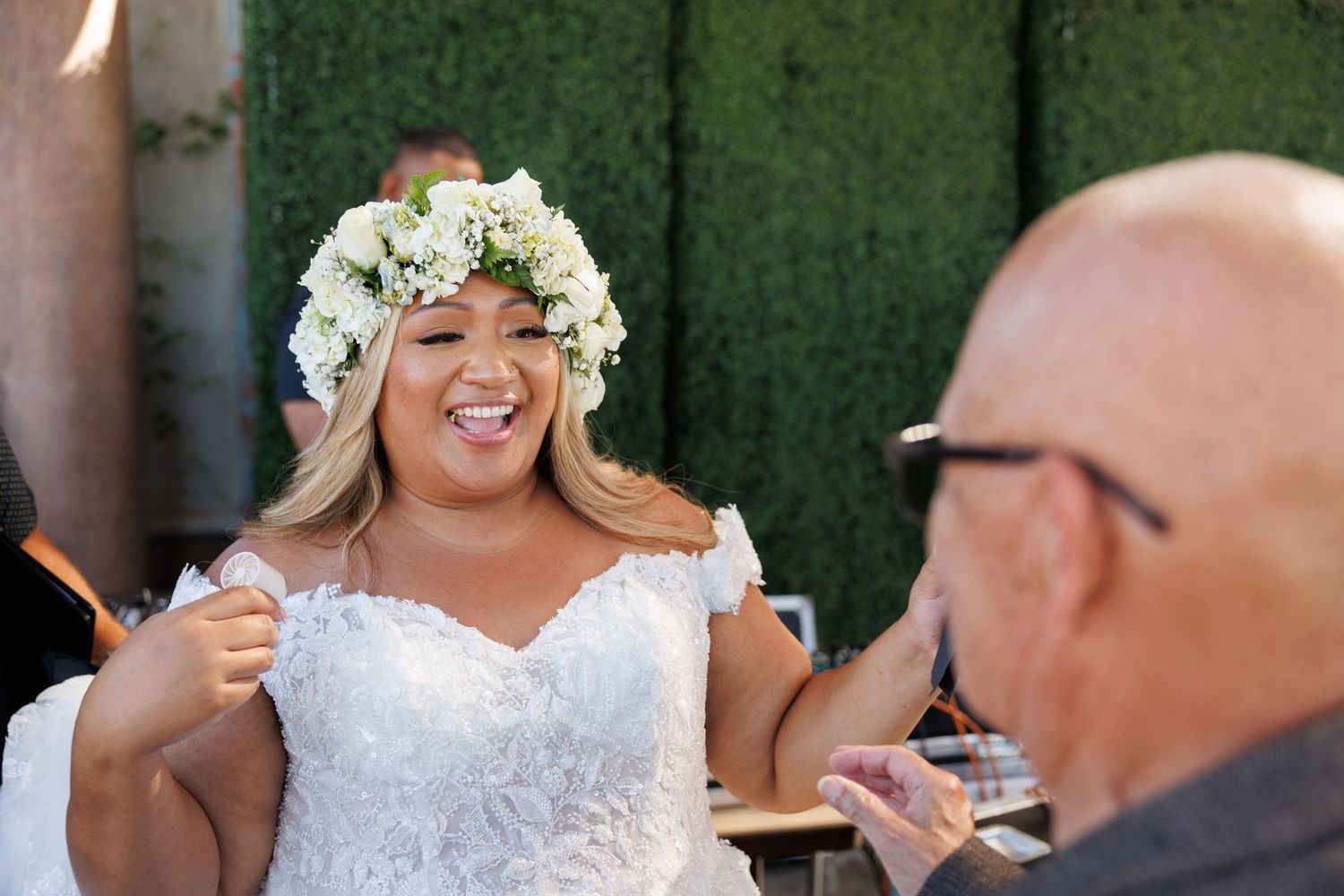 Joyful bride in white lace dress and floral crown laughing during outdoor wedding celebration.