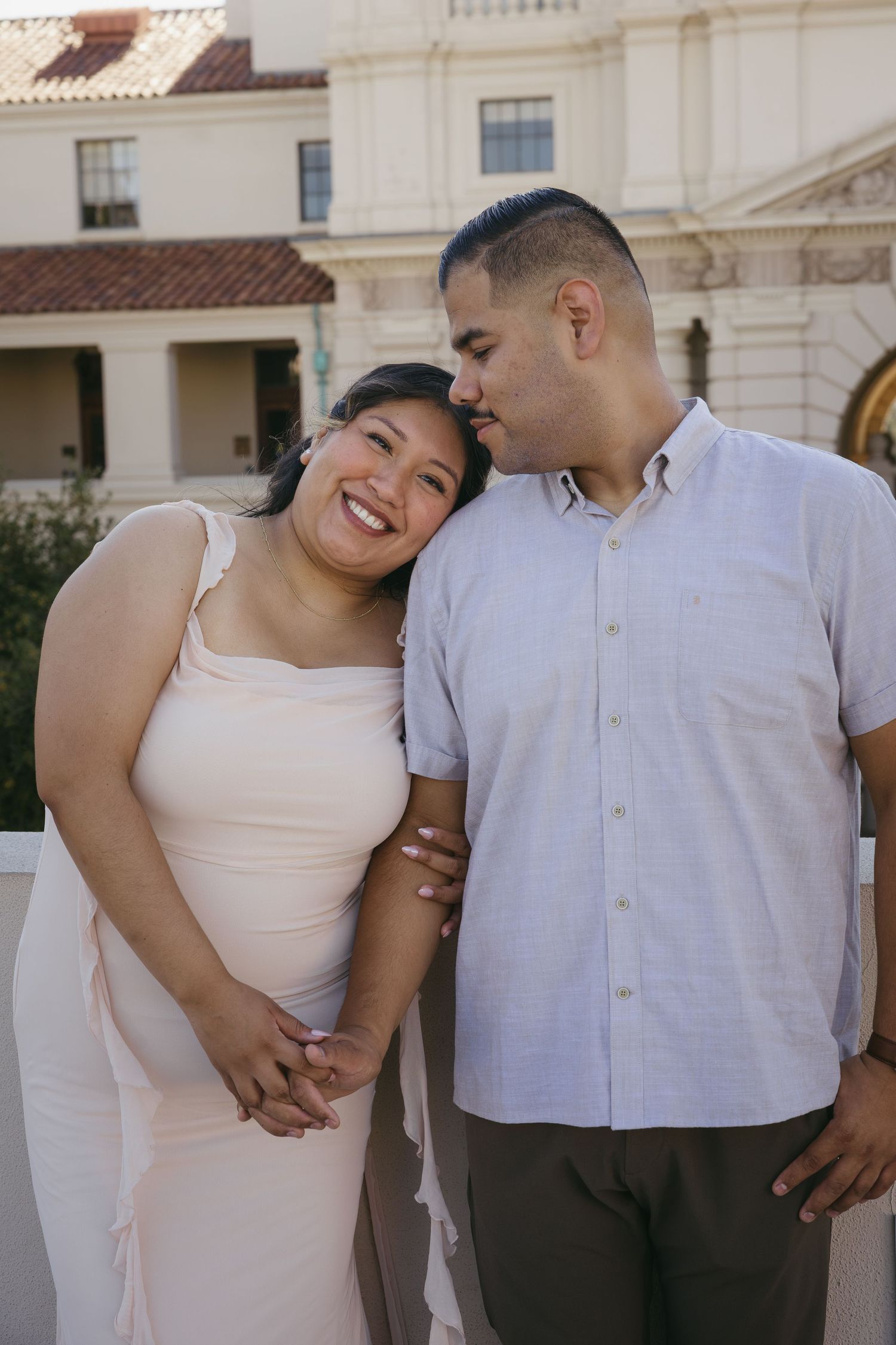A couple shares a tender moment outdoors, man kissing woman's temple while she smiles, near a white building.
