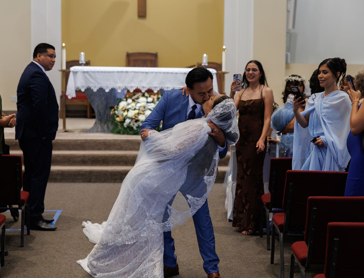 A groom dips his bride in a long lace veil for a kiss in a church aisle as wedding guests watch and take photos.