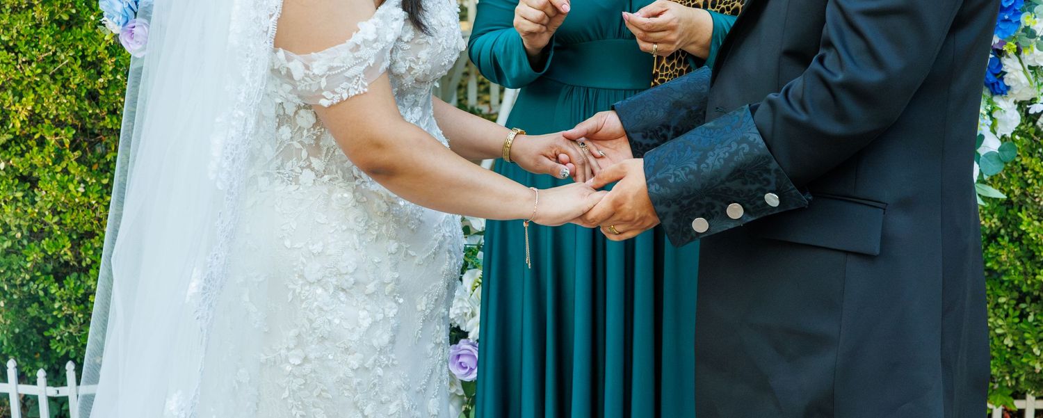 Bride and groom exchanging rings at outdoor wedding ceremony with bridesmaid in teal dress in background.