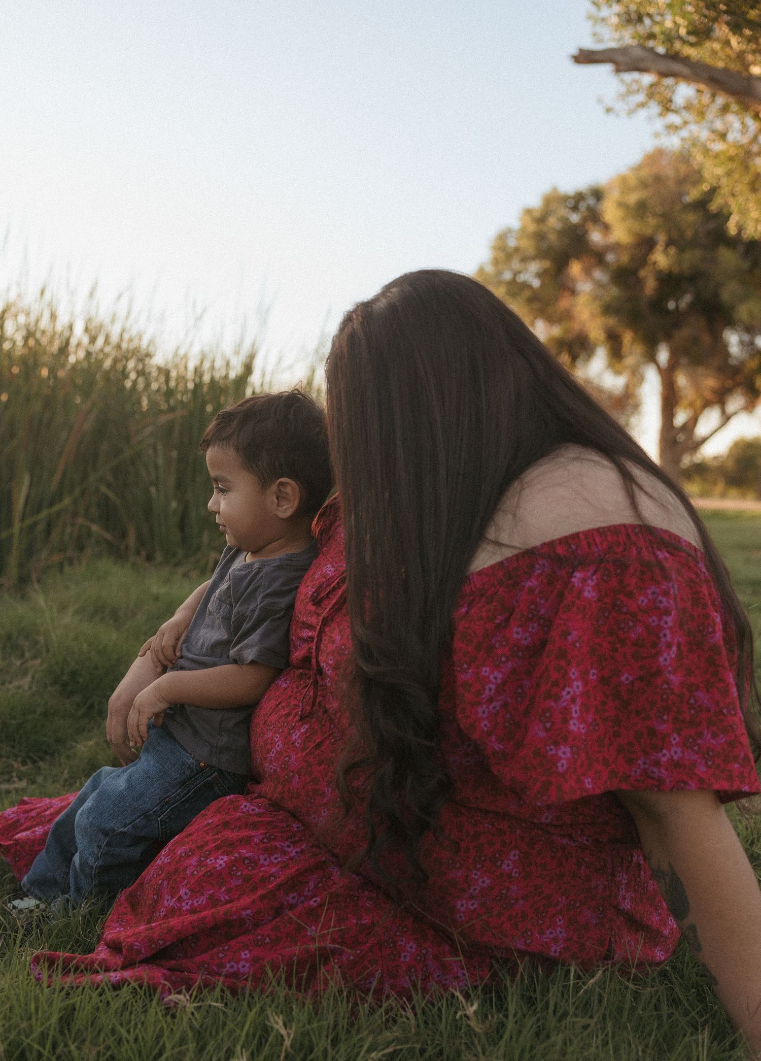 A woman in a red floral off-shoulder dress sits on grass with a toddler boy, both facing away in a serene outdoor setting.