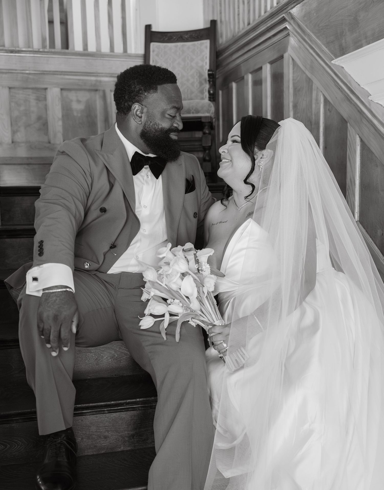 Bride and groom share a loving glance on staircase in black and white wedding photo.