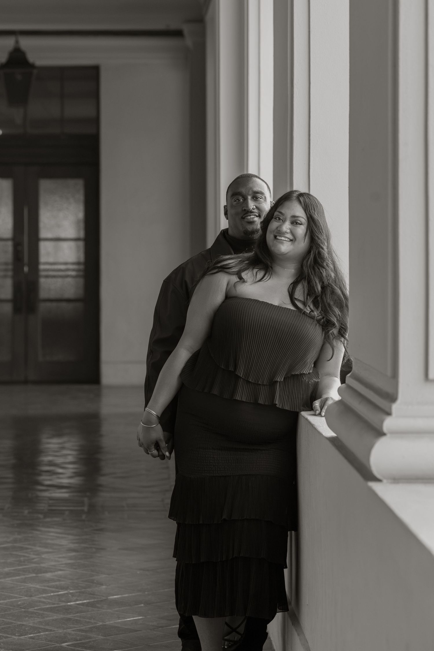 Black and white photo of a couple posing in an elegant hallway, woman in strapless dress leaning against column.