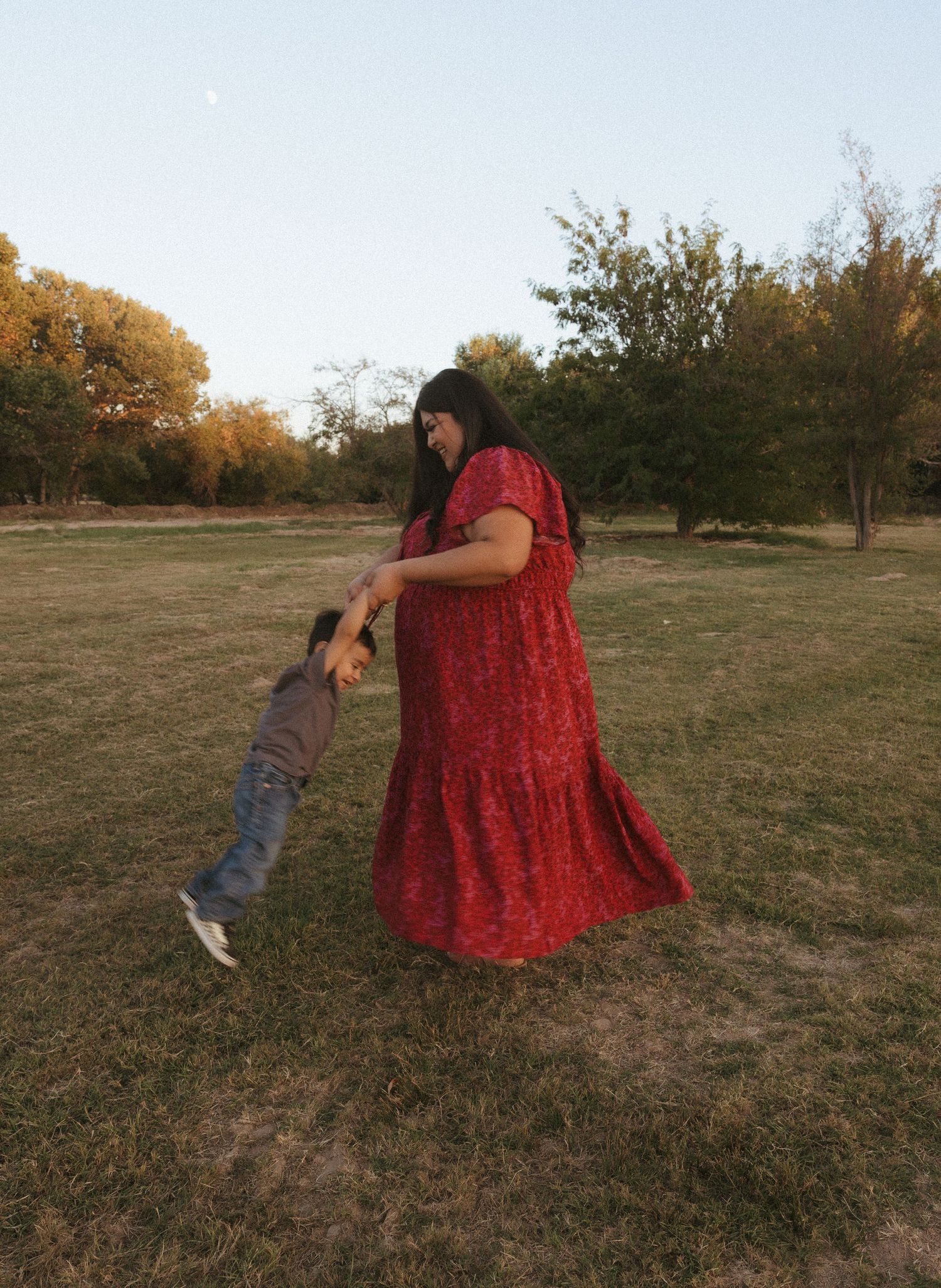 A mother in a long red dress playfully swings her toddler by the hands in a grassy park.
