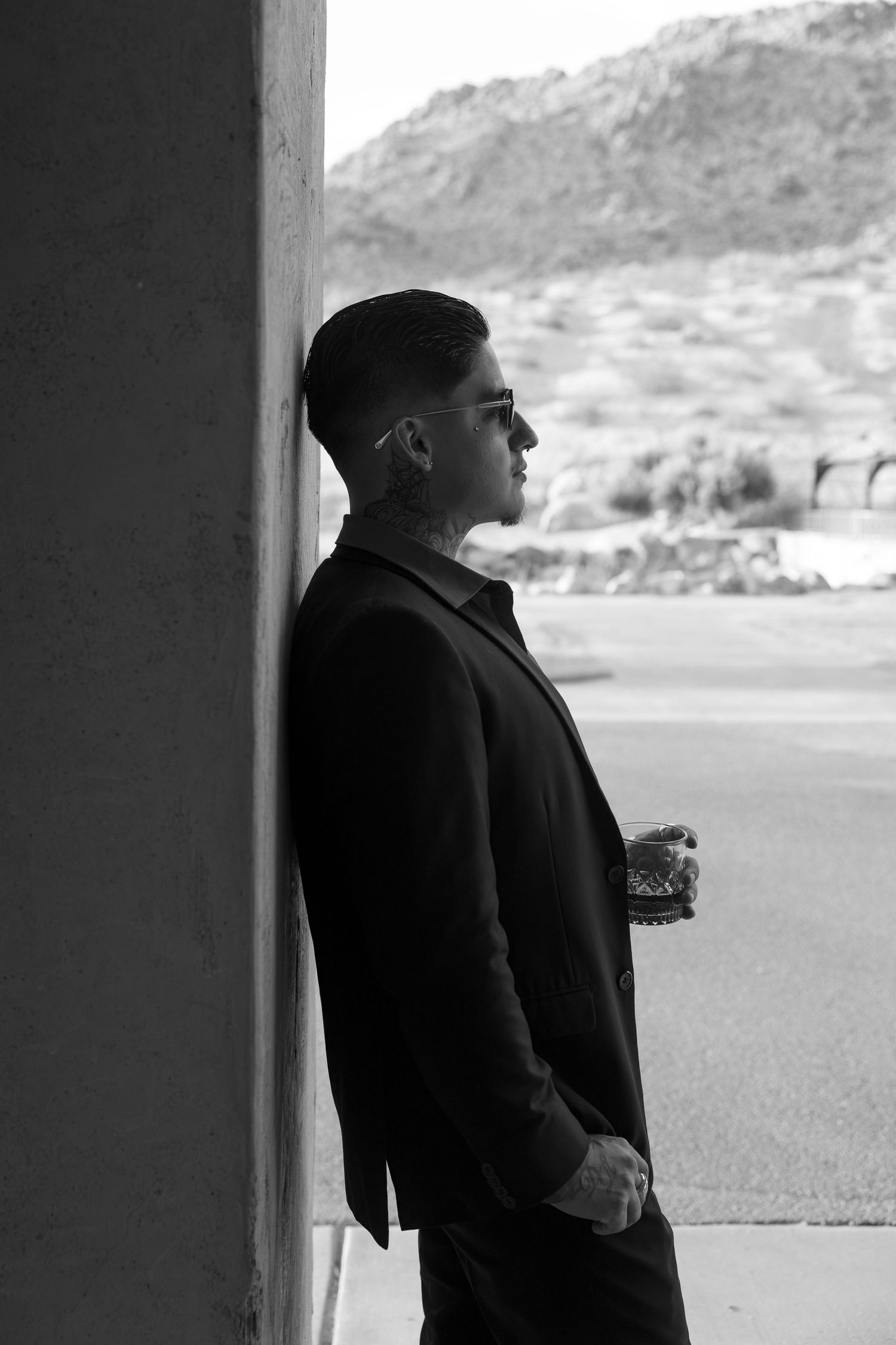 Tattooed man in black jacket leaning against wall with desert landscape in black and white photo.