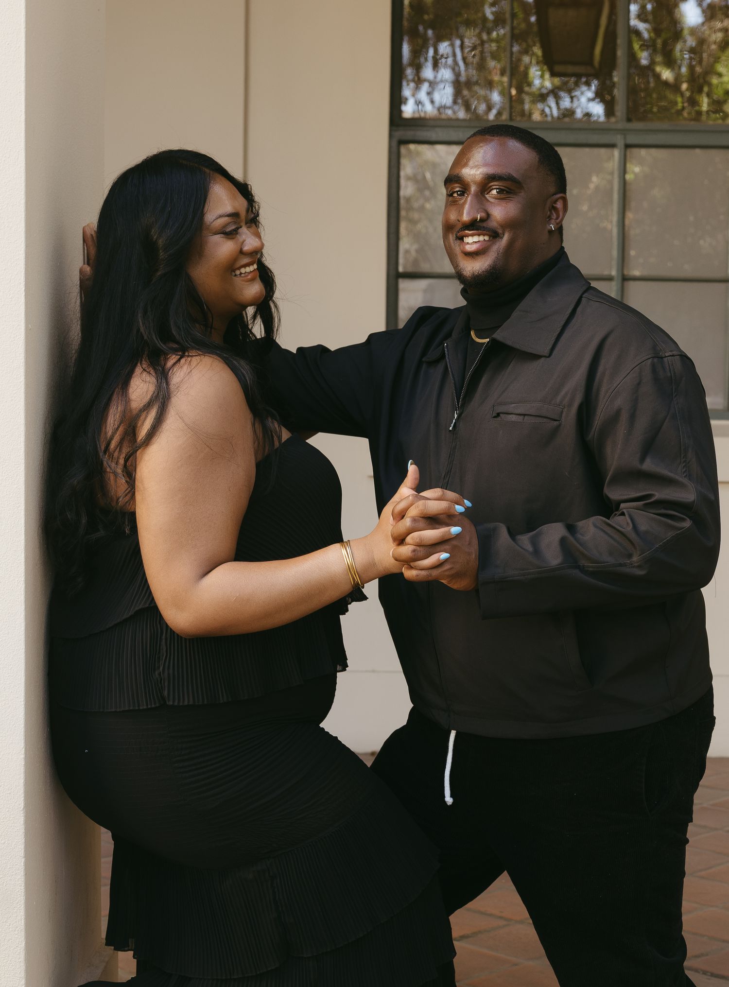 A joyful couple dressed in black laughing and holding hands against a white pillar outdoors.