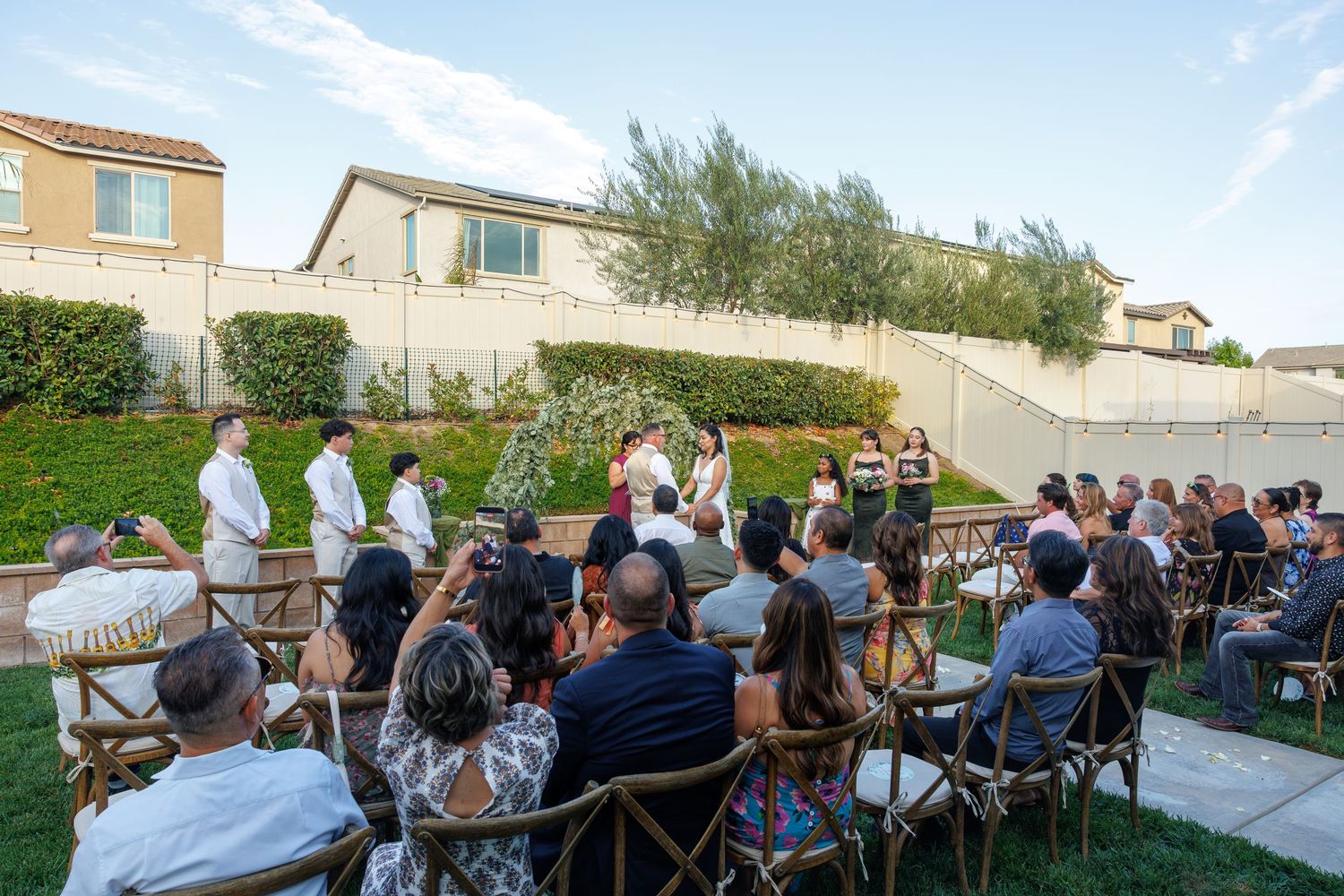 Outdoor backyard wedding ceremony with guests seated on wooden chairs facing the altar under a clear blue sky.