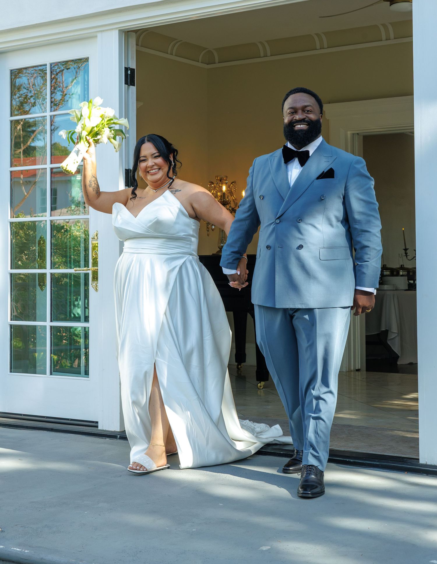 Joyful newlyweds exit venue, bride in white gown holding bouquet, groom in light blue suit, holding hands.