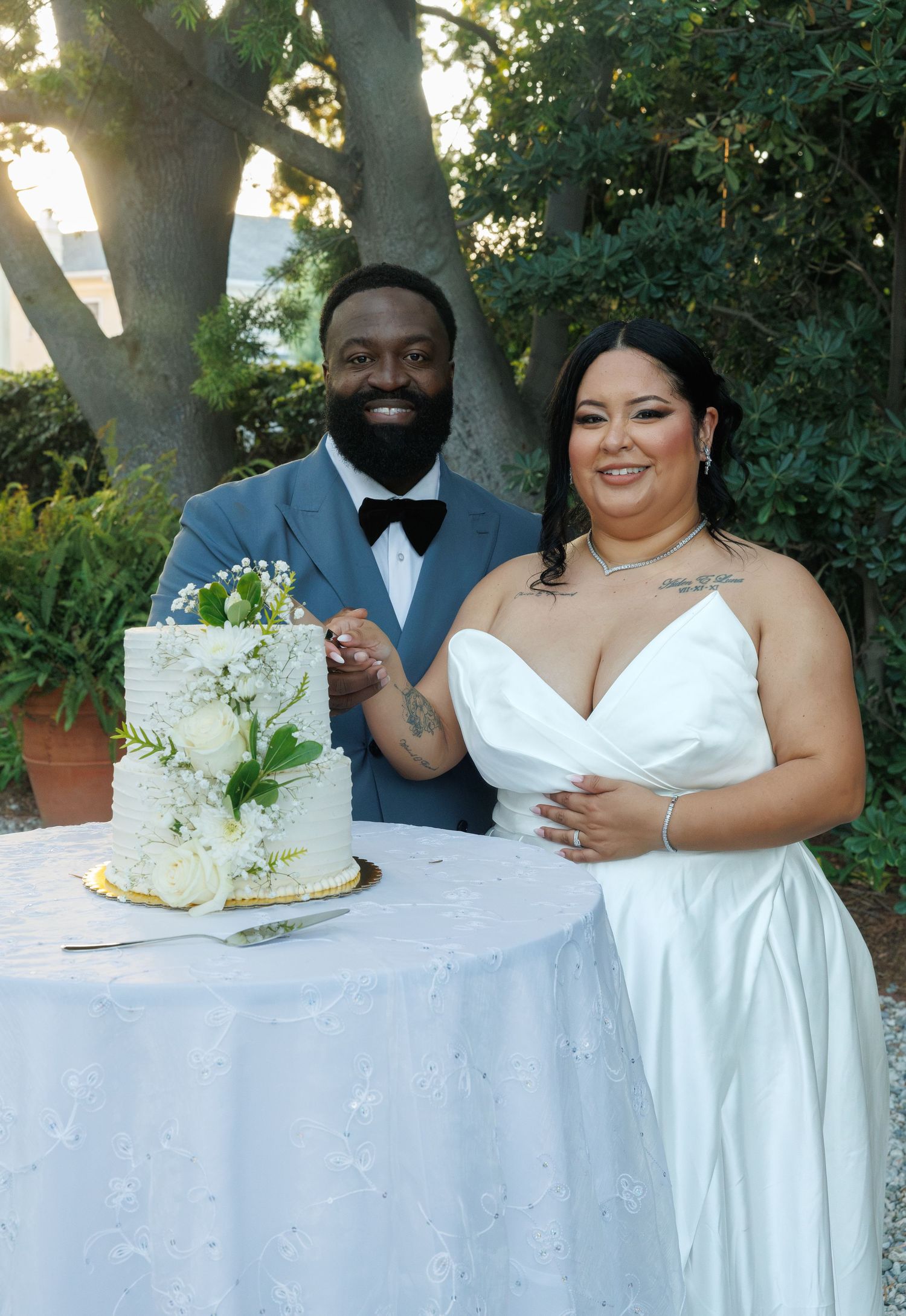 Smiling bride and groom cutting white floral wedding cake at outdoor garden reception.