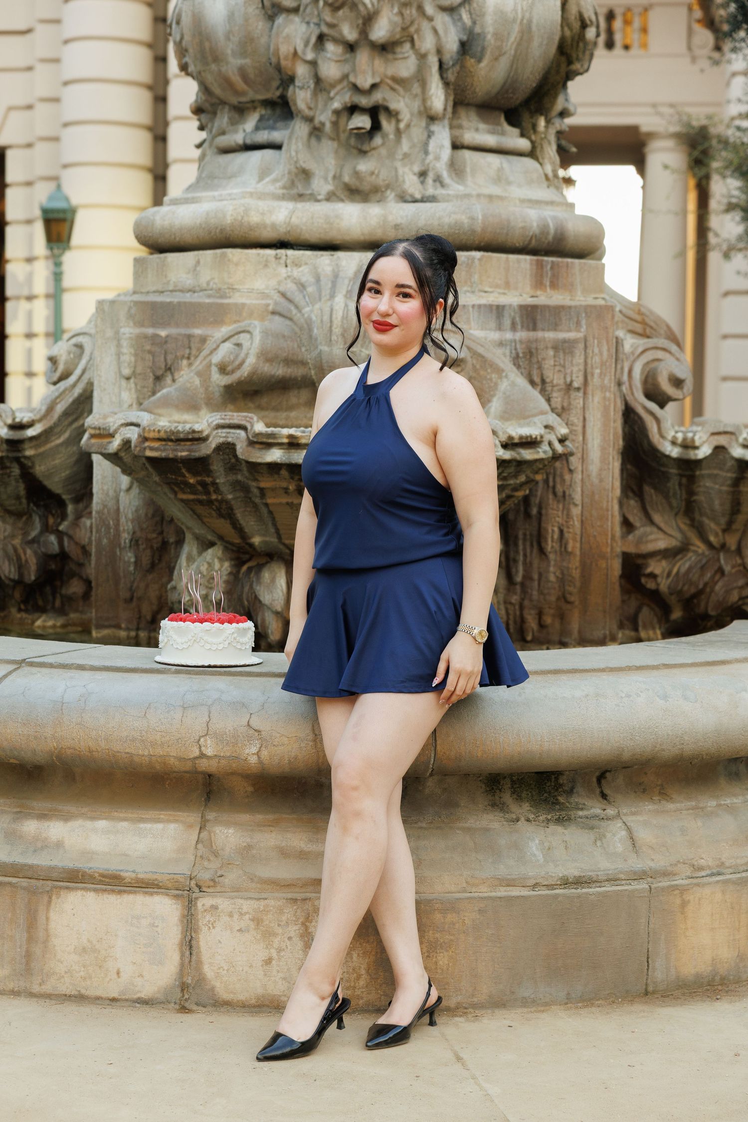 Woman in navy blue halter romper poses by ornate stone fountain with birthday cake, wearing black heels.